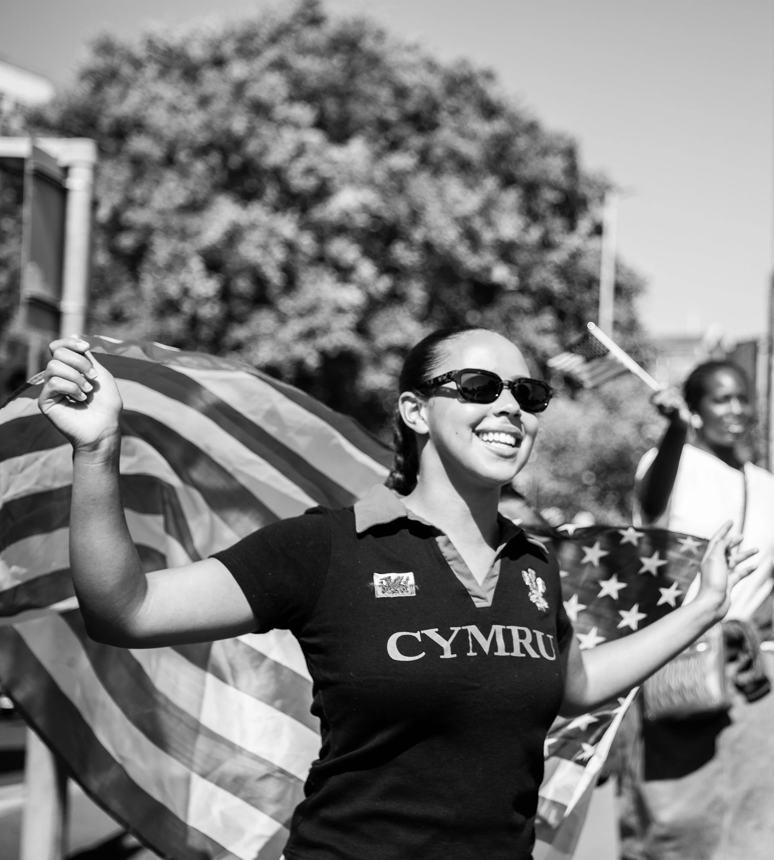 A woman smiling and wearing sunglasses, holding an American flag and a Cymru shirt, participating in a parade or celebration outdoors with trees in the background.
