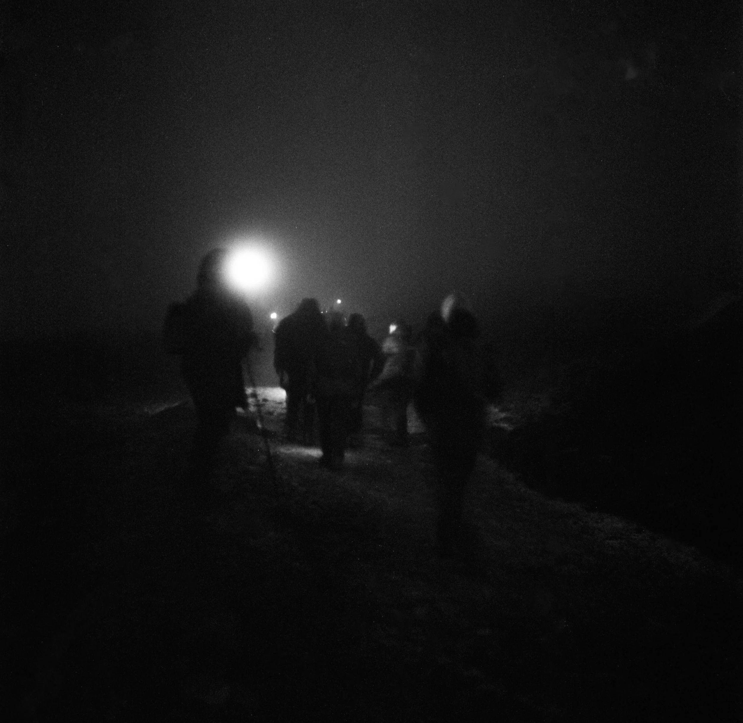 A group of people walking along a dark, foggy beach at night with a bright moon or light source in the distance.
