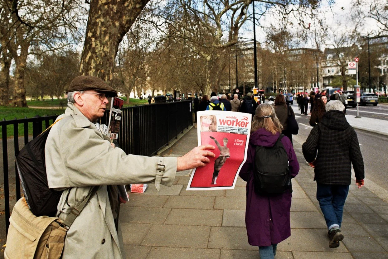Man selling newspapers on a city sidewalk, with pedestrians walking past and trees in the background.