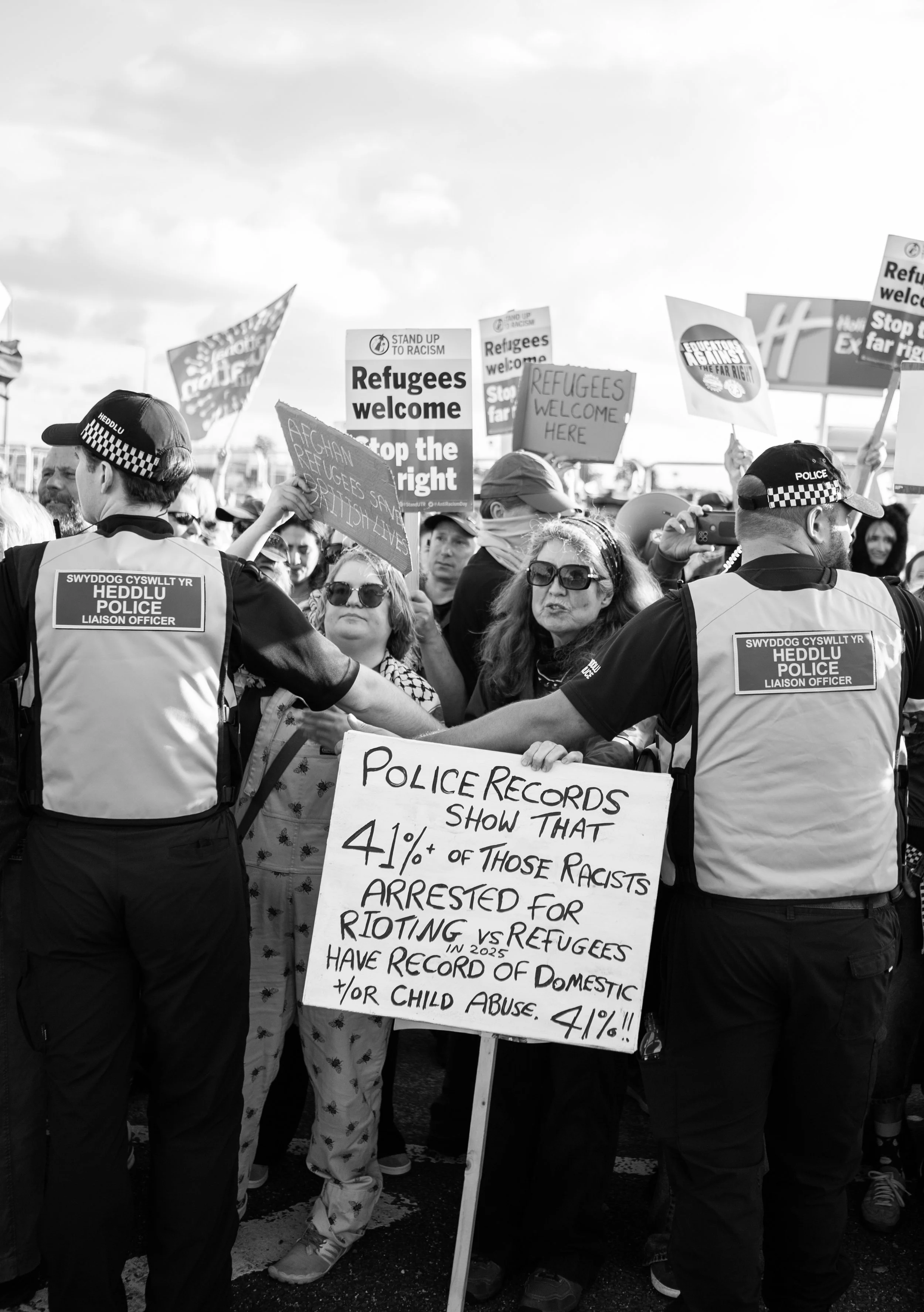 Black and white photo of a protest with people holding signs, and police officers holding a controversial sign in the foreground that states police records show a percentage of racist arrests, with many signs in the background supporting refugees and