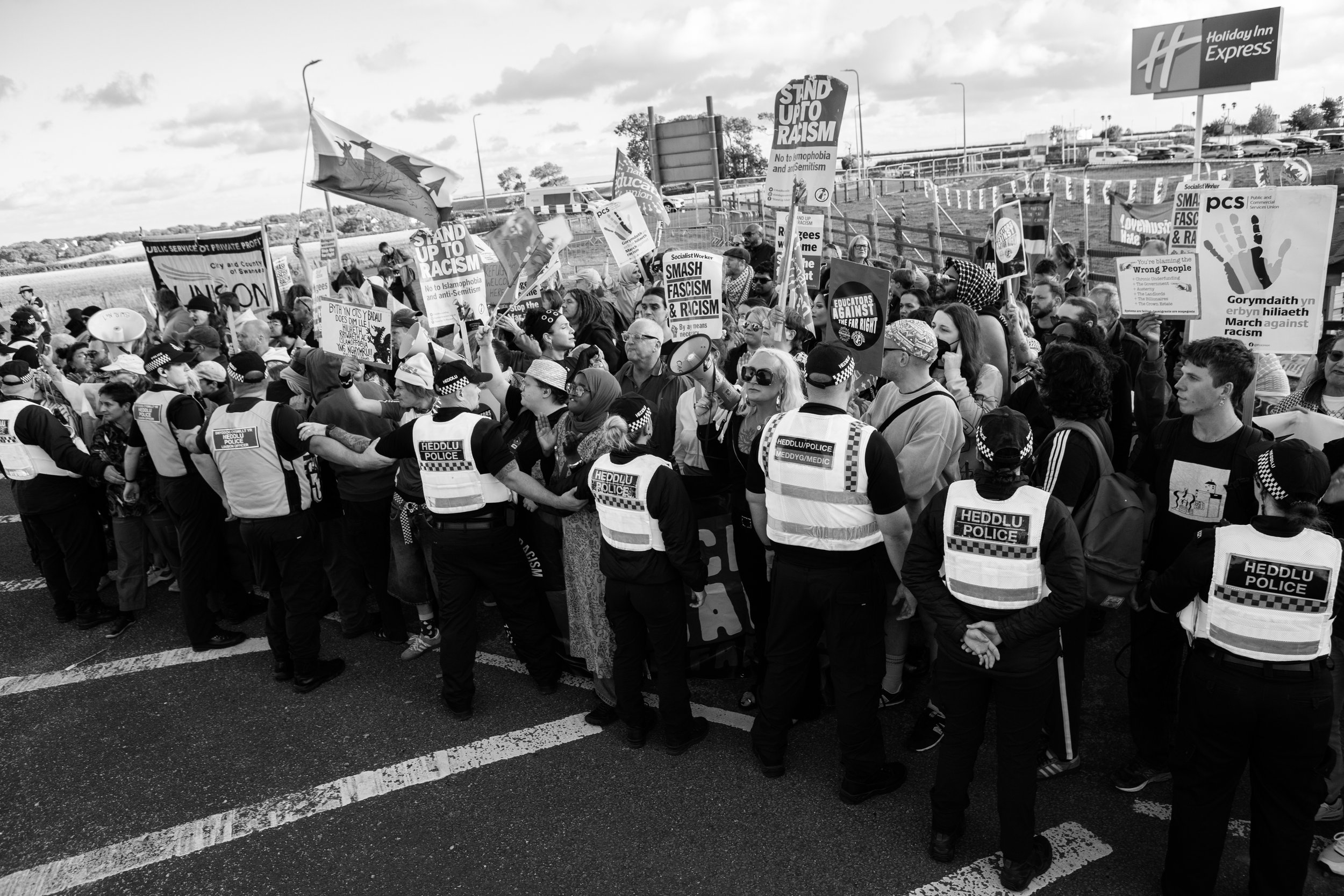 Crowd of protesters holding signs and banners, gathered behind police officers in a line during a demonstration against racism and fascism, with police in the foreground.