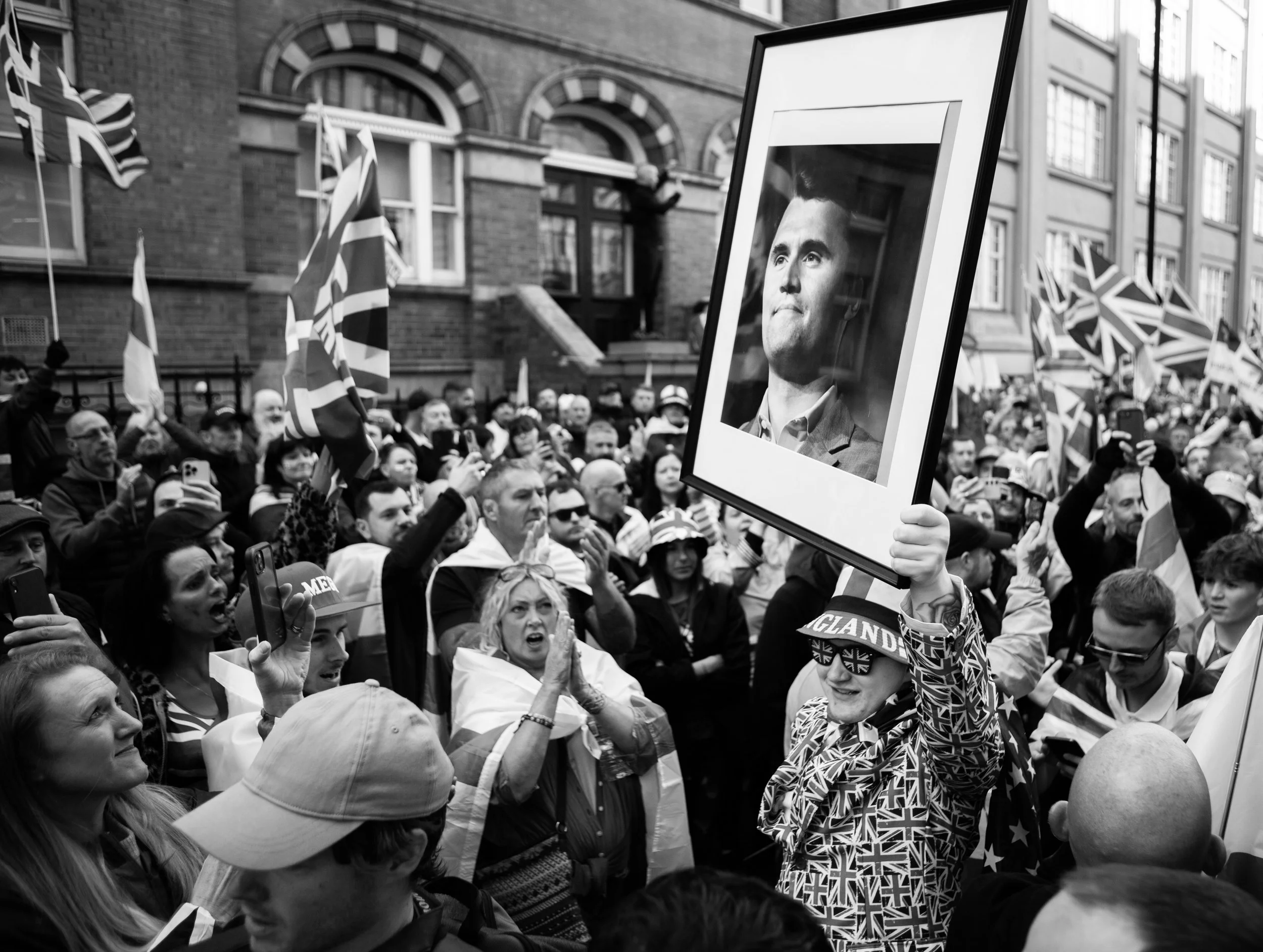 Crowd of people at a protest or rally, with flags and banners, one person holding a framed black-and-white portrait of Charlie Kirk.