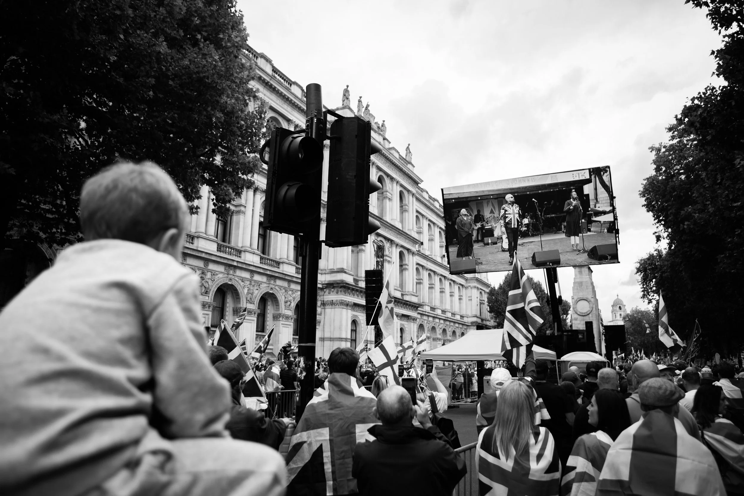 Crowd of people attending a rally or protest, many holding Union Jack flags, watching a stage with performers on a large screen, in front of a historic building with ornate architecture, on a cloudy day.