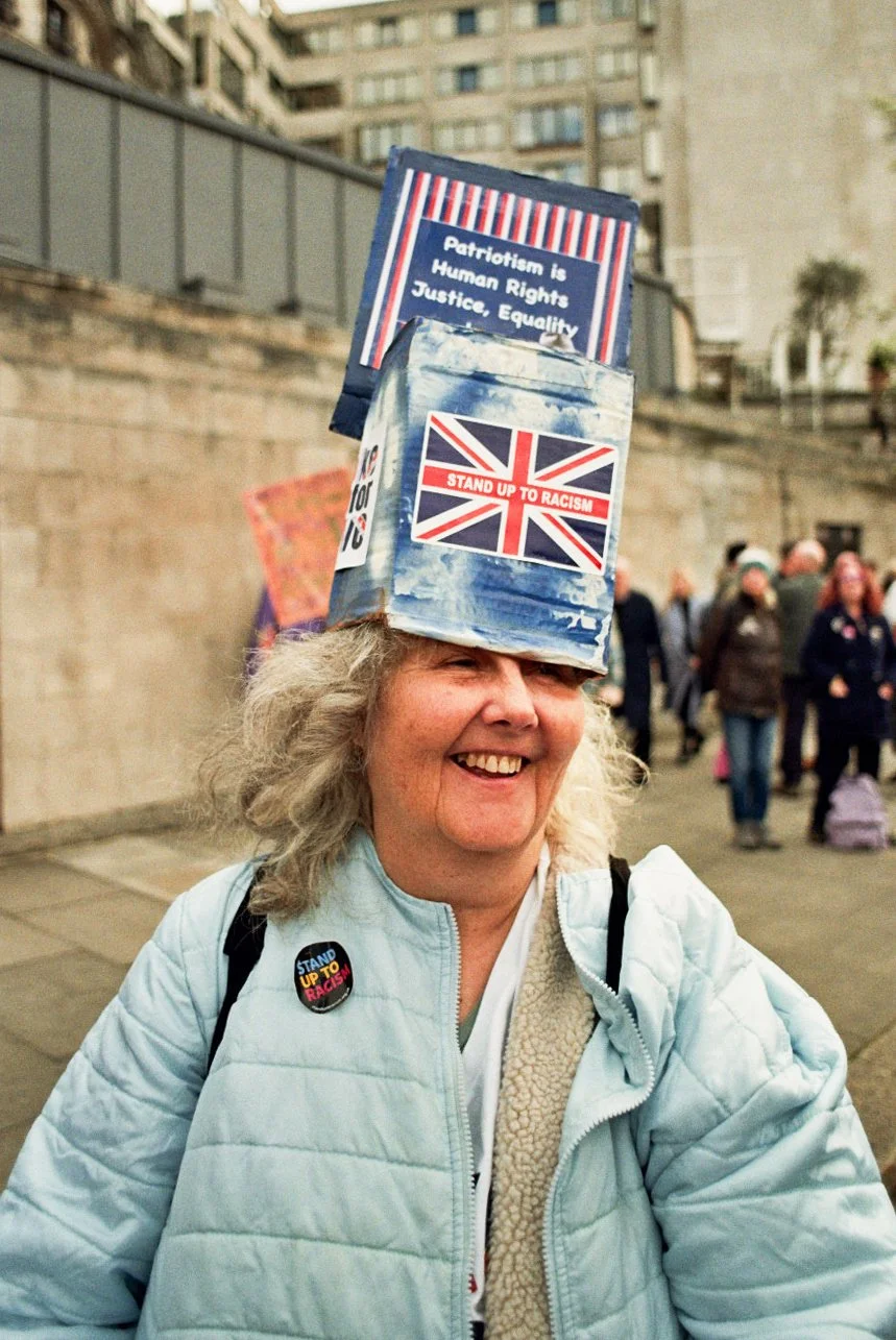 A woman with gray hair smiling at an outdoor event, wearing a white jacket with a 'Stand Up to Racism' button, and a cardboard hat decorated with British flags and signs advocating for patriotism, human rights, justice, equality, and standing up to r