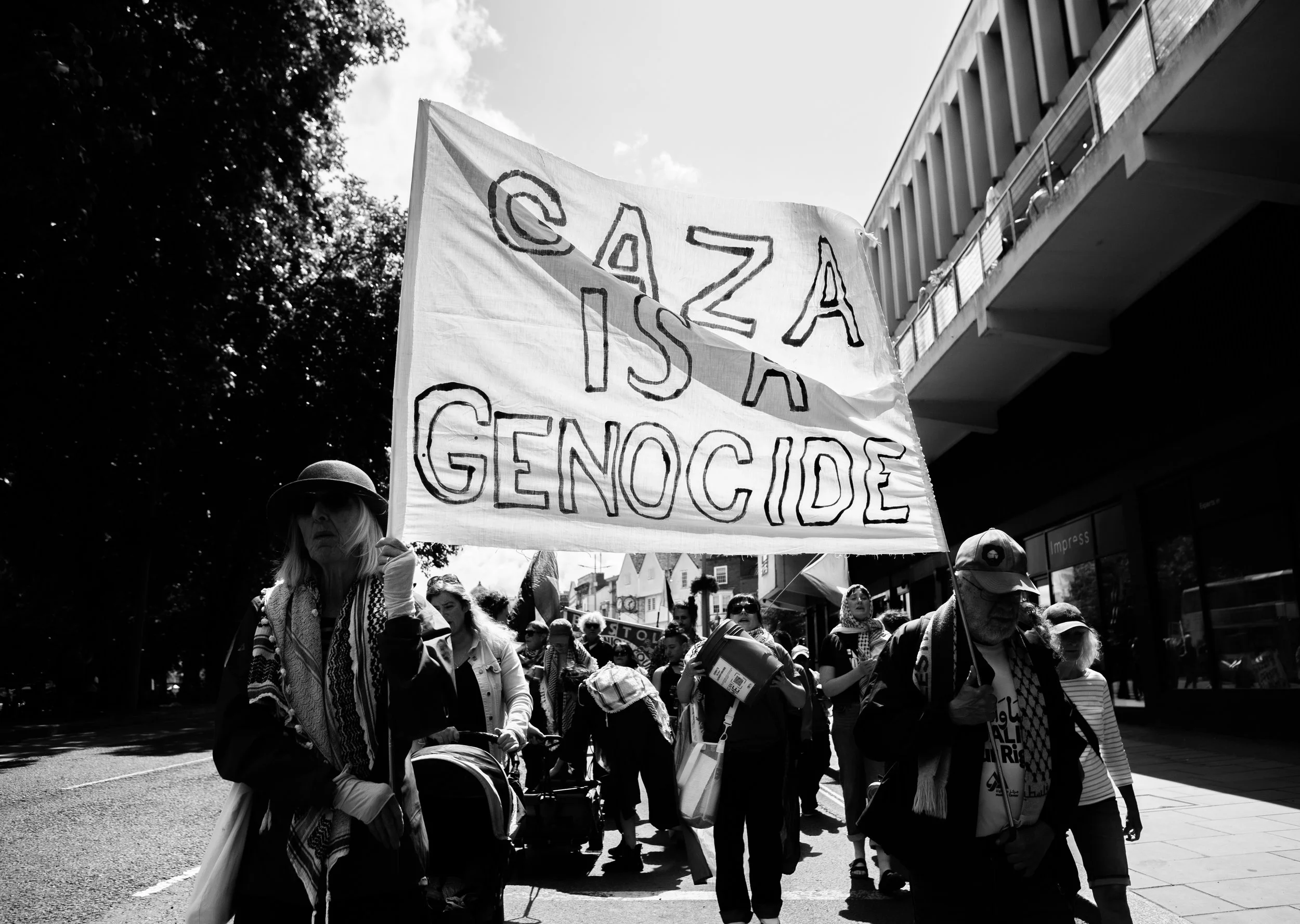 People protesting in a march holding a large sign that reads 'Caza is a genocide' in black and white.