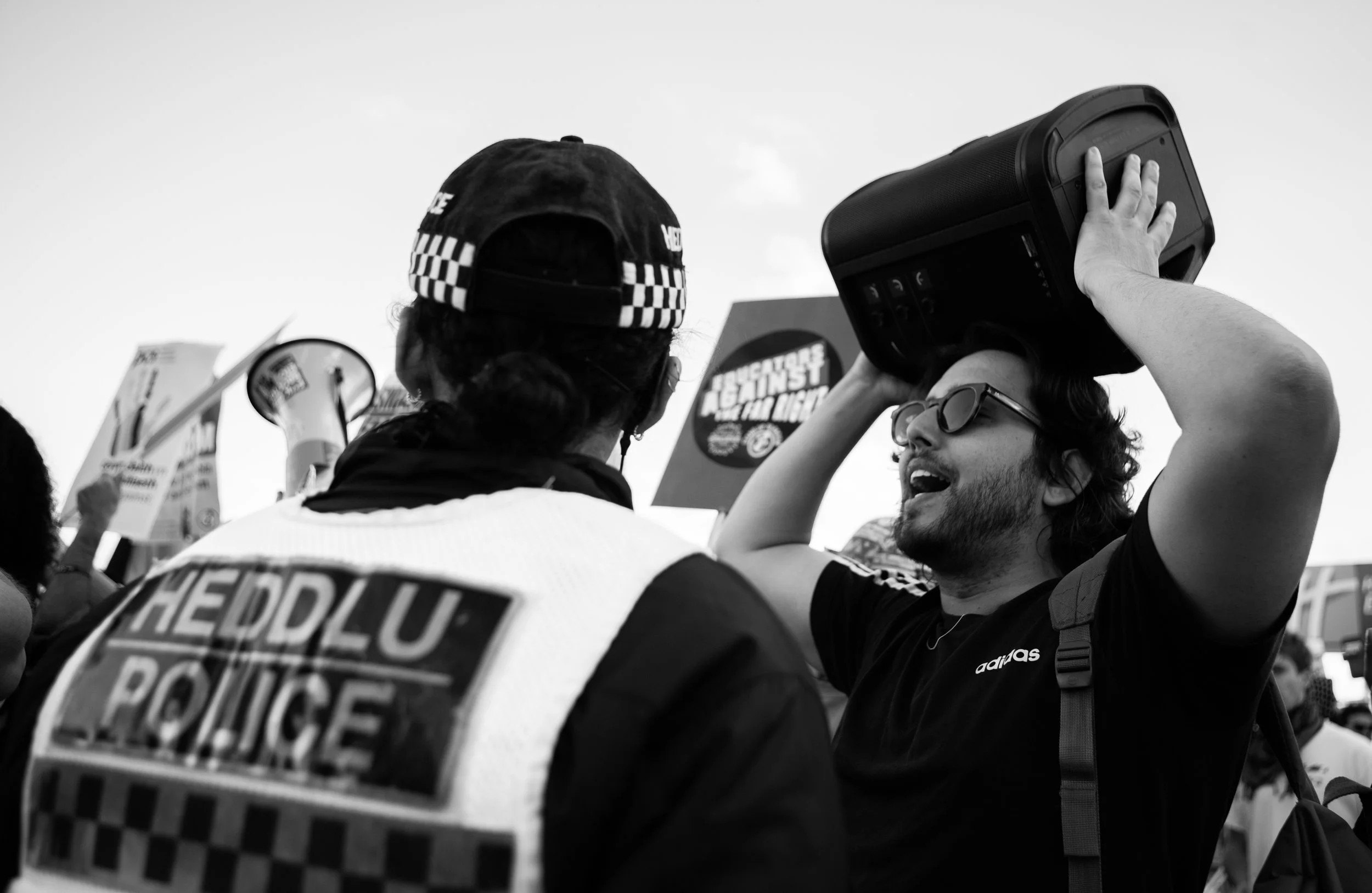 A man with sunglasses holding a speaker talks to a police officer at a protest or rally, with signs and banners visible in the background.