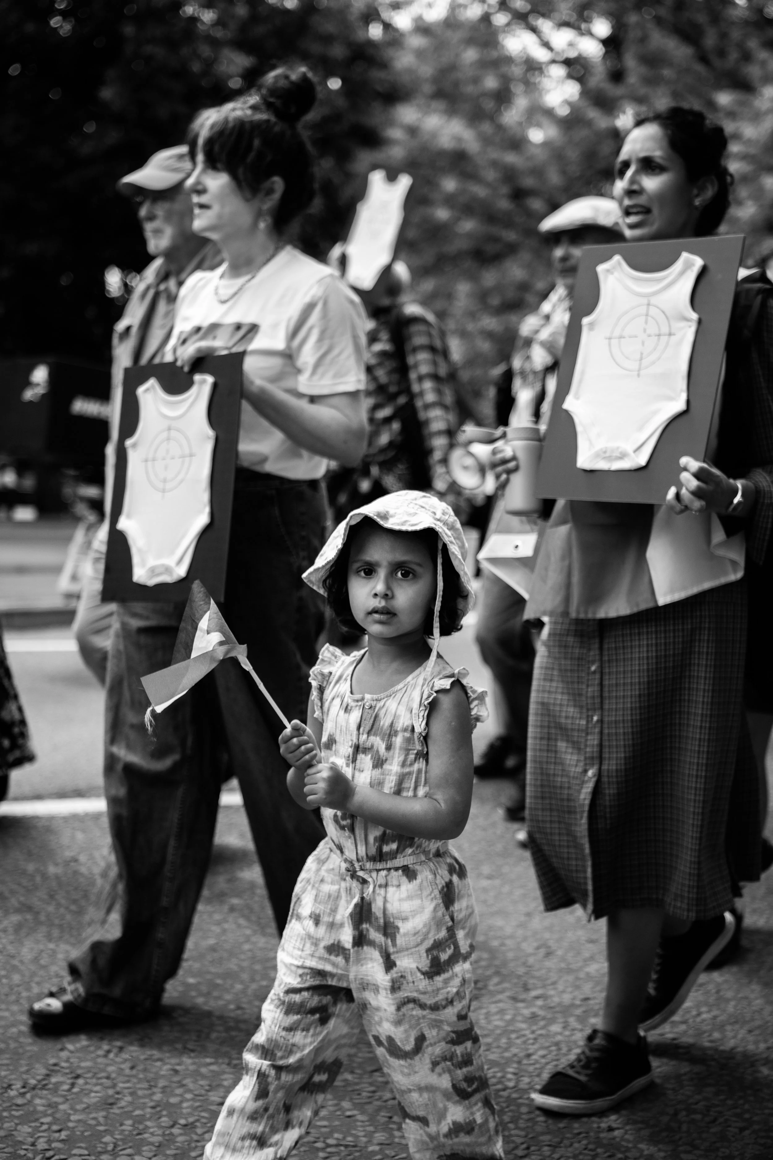 A young girl holding a small flag participating in a protest or march, with adult women holding signs with target symbols on them in the background.