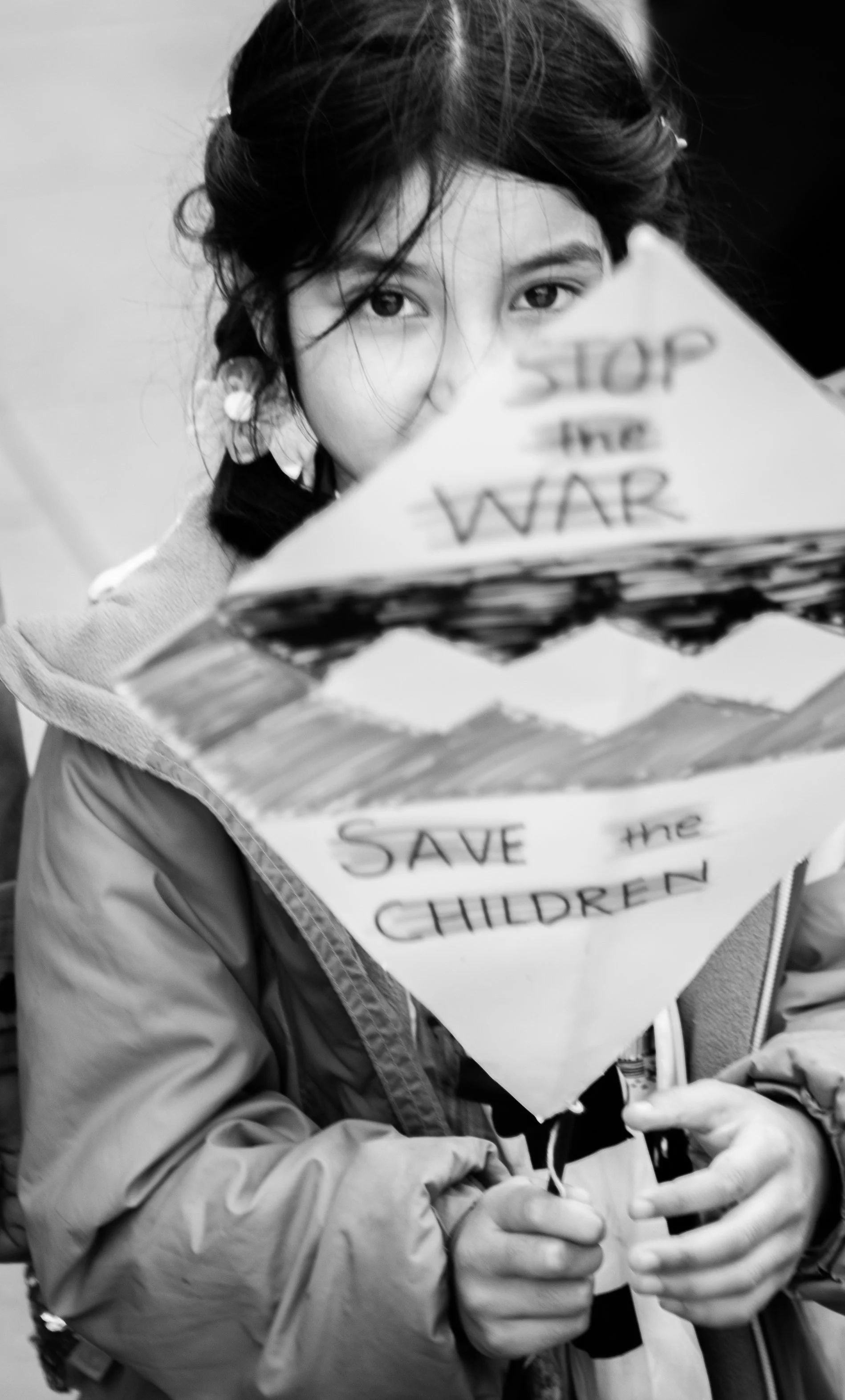 A young girl with dark hair, wearing a jacket, holding a homemade protest sign that reads 'Stop the WAR' and 'Save the CHILDREN', with a serious expression.