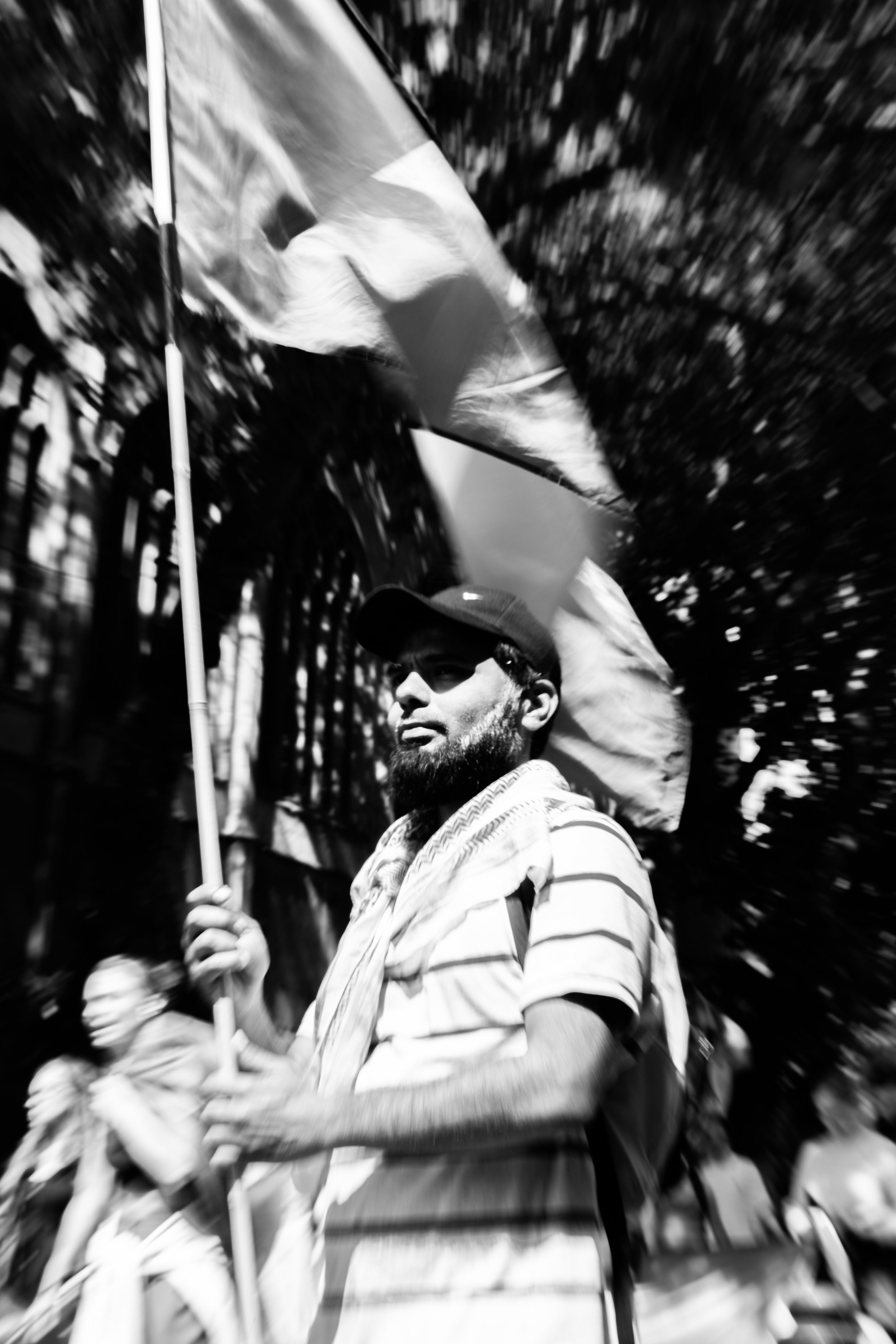 A man with a beard holding a flag during a protest or rally, with a blurred background of trees and other people.