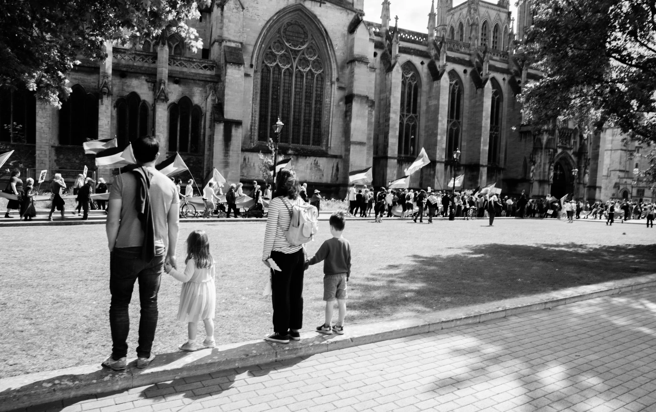 A black and white photo of a large group of people gathered in front of a historic Gothic cathedral, with people waving flags and some participating in a procession, while a family with a man, woman, and two children watches from the sidewalk.
