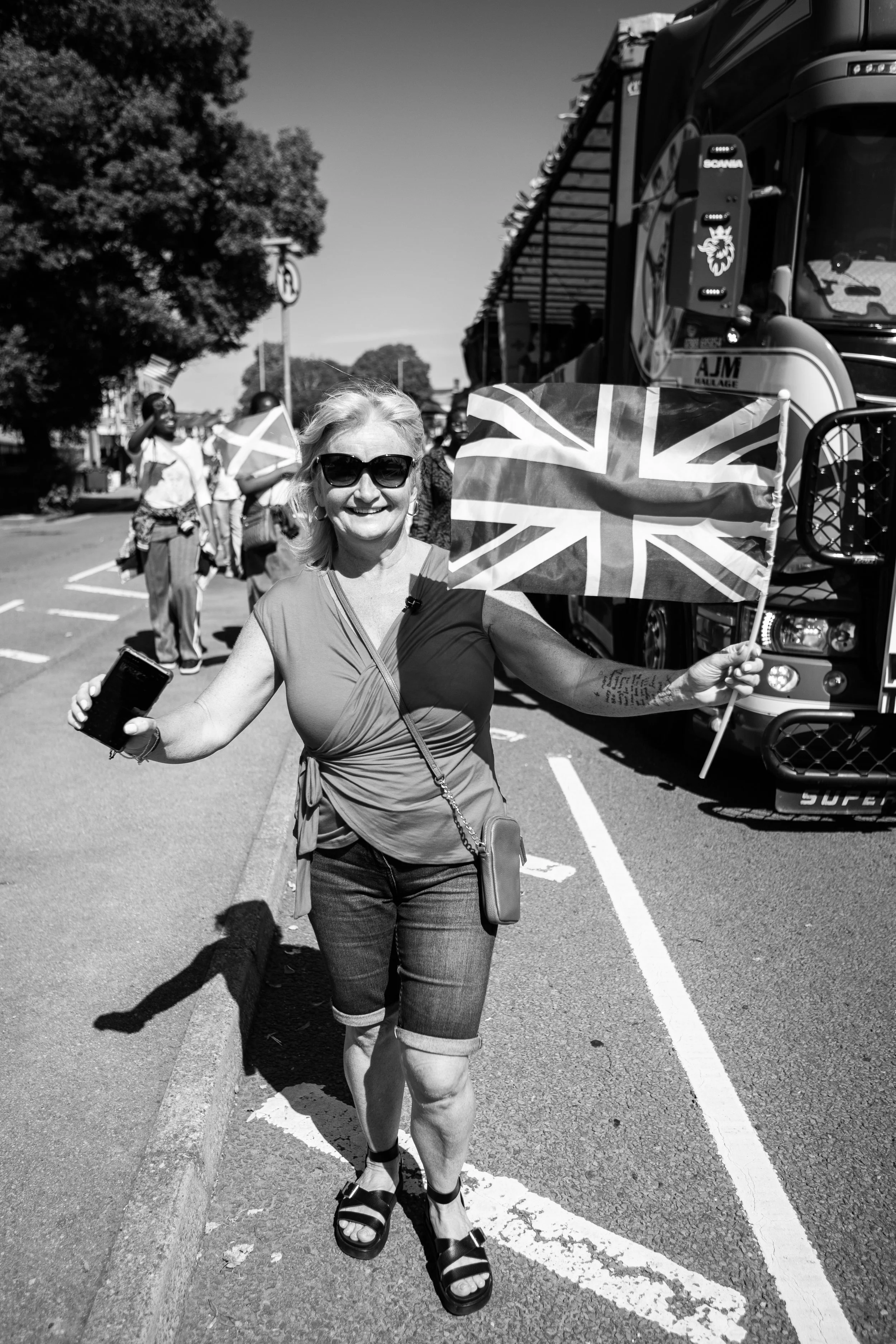 A smiling woman wearing sunglasses, holding a mobile phone and a Union Jack flag, standing on a street during a sunny day with a large truck and other people in the background.