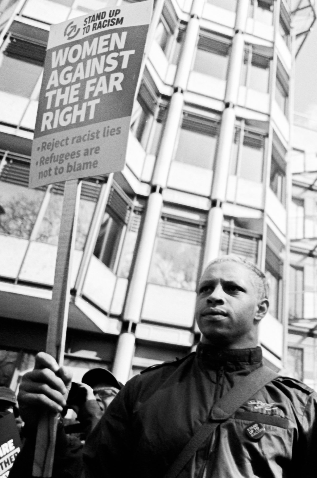 A Black man holding a protest sign that reads "Women Against The Far Right," with smaller text saying "Reject racist lies" and "Refugees are not to blame," at a rally during a demonstration against racism, with a modern building in the background.