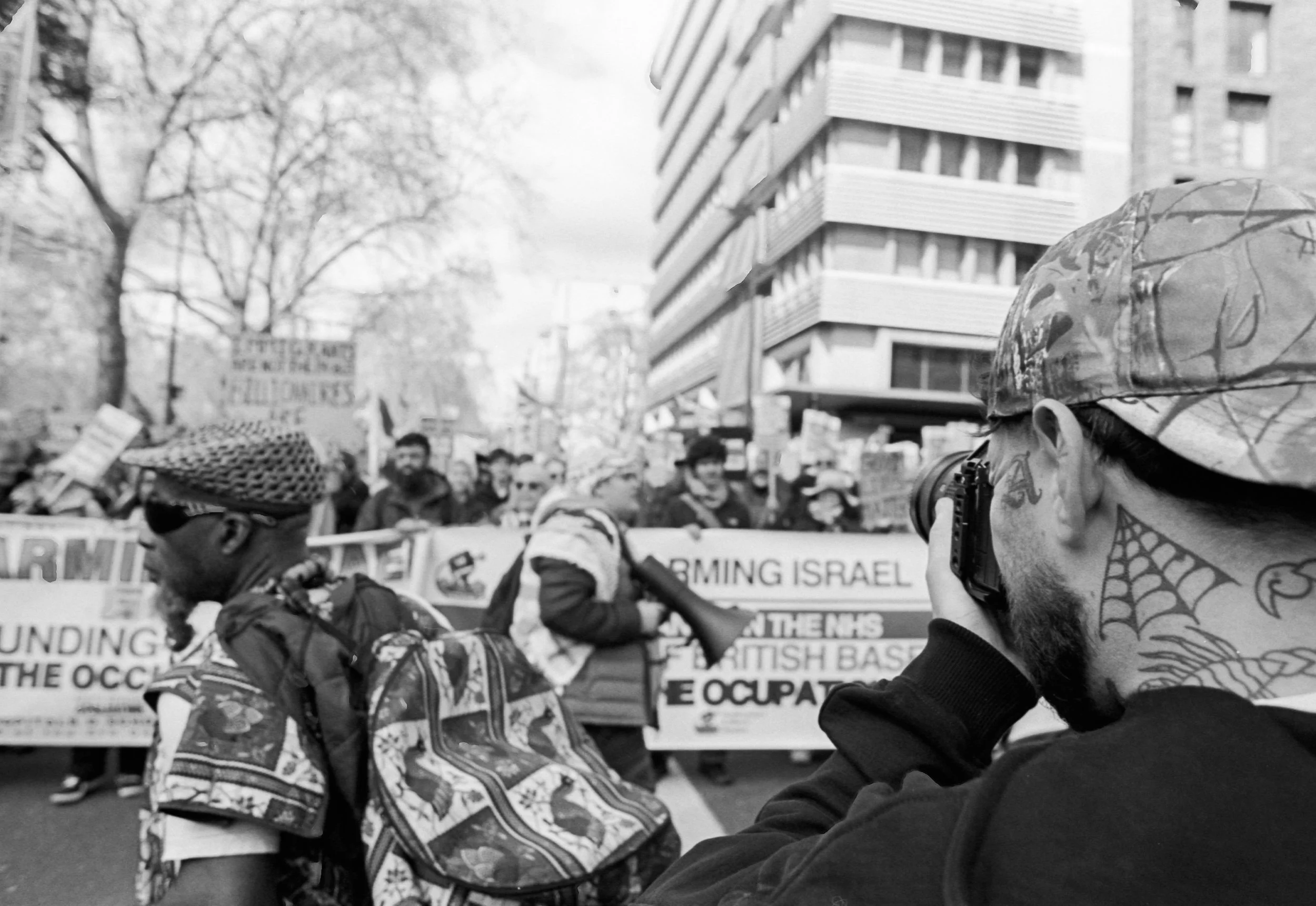 A black and white photo of a protest with multiple people holding signs. In the foreground, a man with a beard and tattoos, wearing a camouflage cap, is taking a photo with a camera. The crowd behind him includes individuals with backpacks and signs 