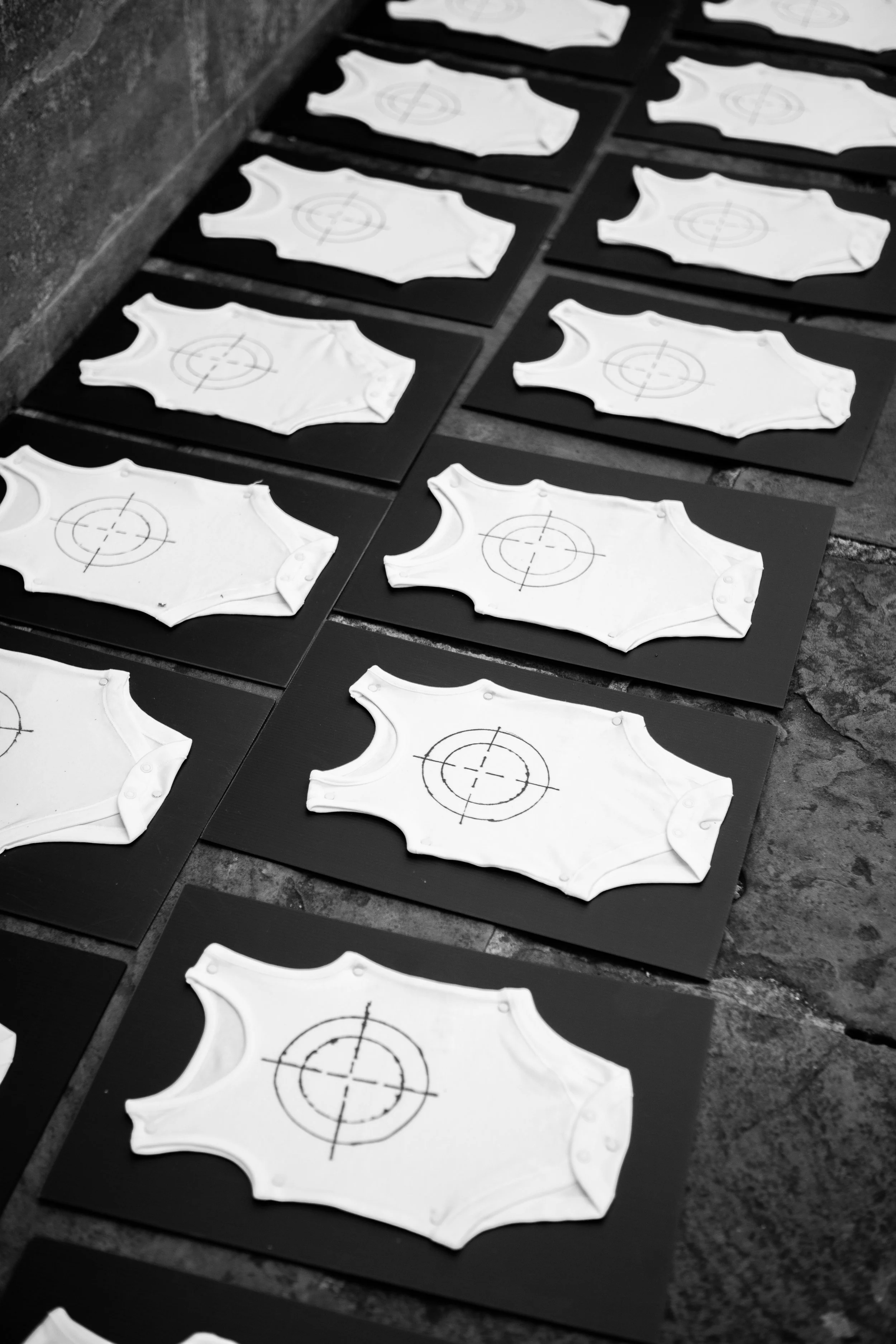 Multiple white clay or ceramic tank tops with target symbols drawn on them, arranged in a grid on black boards, placed on a stone floor.