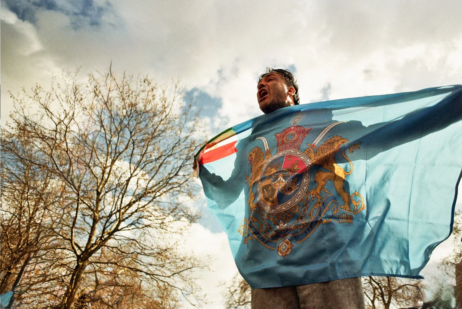 A man holding a flag with a coat of arms, outdoors with bare trees and cloudy sky in the background.