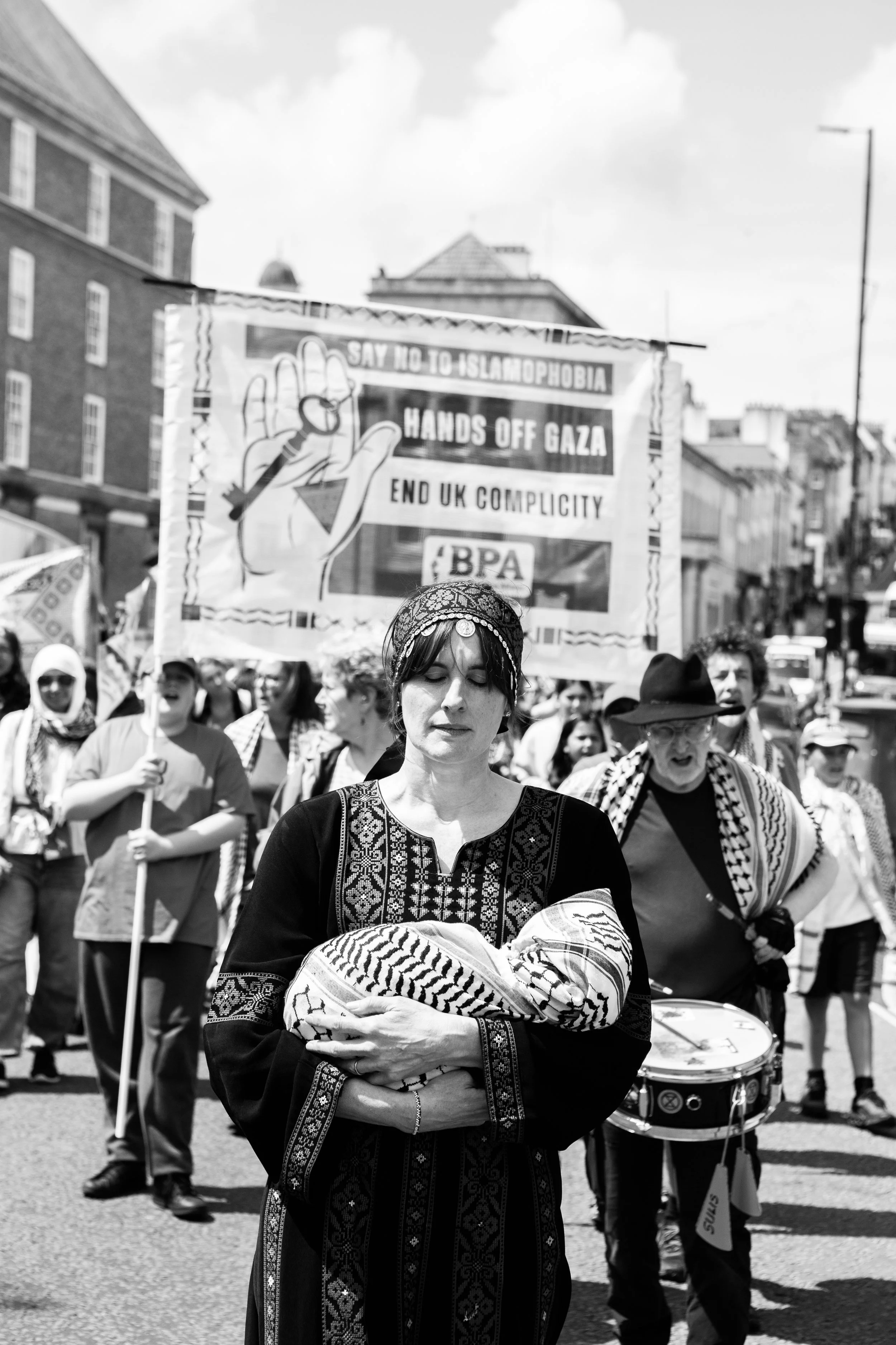 A woman in traditional Palestinian attire holding a baby wrapped in a keffiyeh scarf, with a protest march and a large banner in the background that reads 'Say no to Islamophobia, Hands off Gaza, End UK complicity, BPA.'