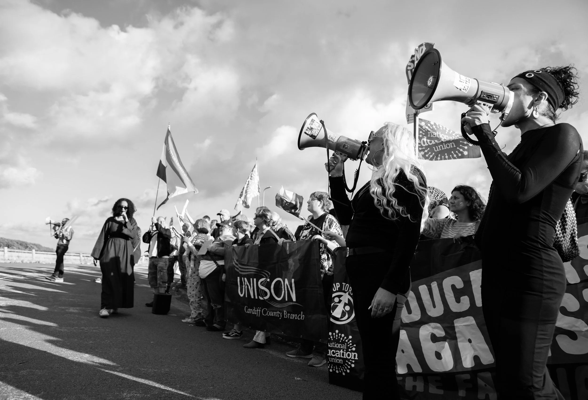 Protesters holding banners and flags outdoors, some speaking into megaphones, participating in a rally or demonstration on a partly cloudy day.