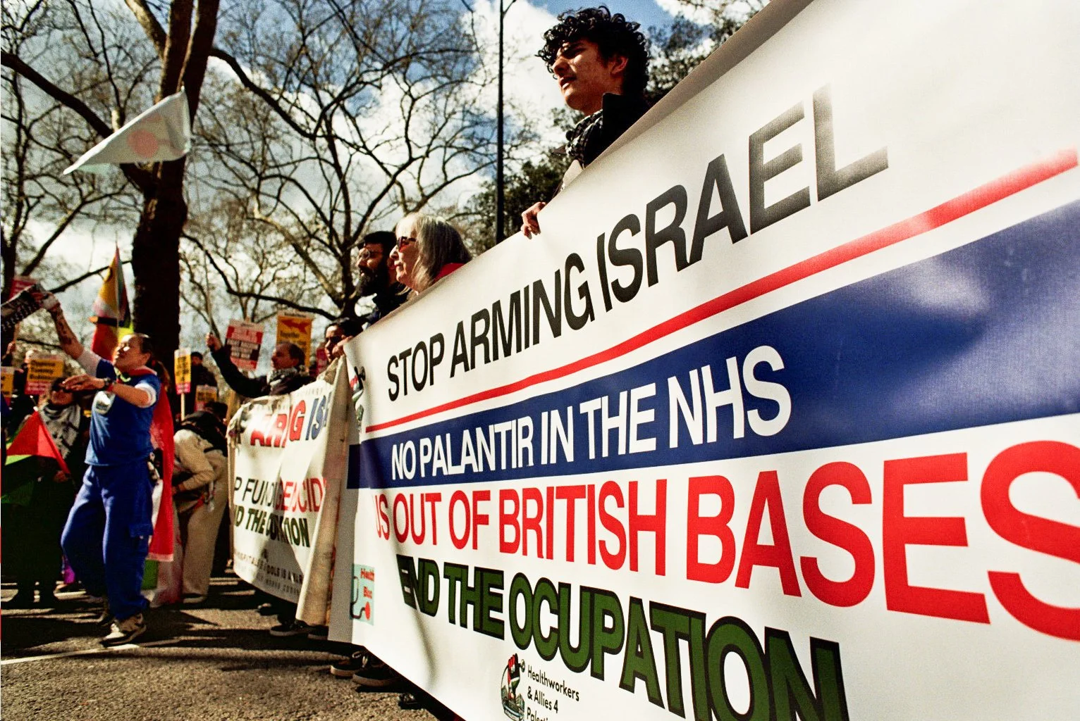 Protesters holding a large banner that says 'Stop Arming Israel, No Palantir in the NHS, Out of British bases, End the occupation' during a march on a street with leafless trees.