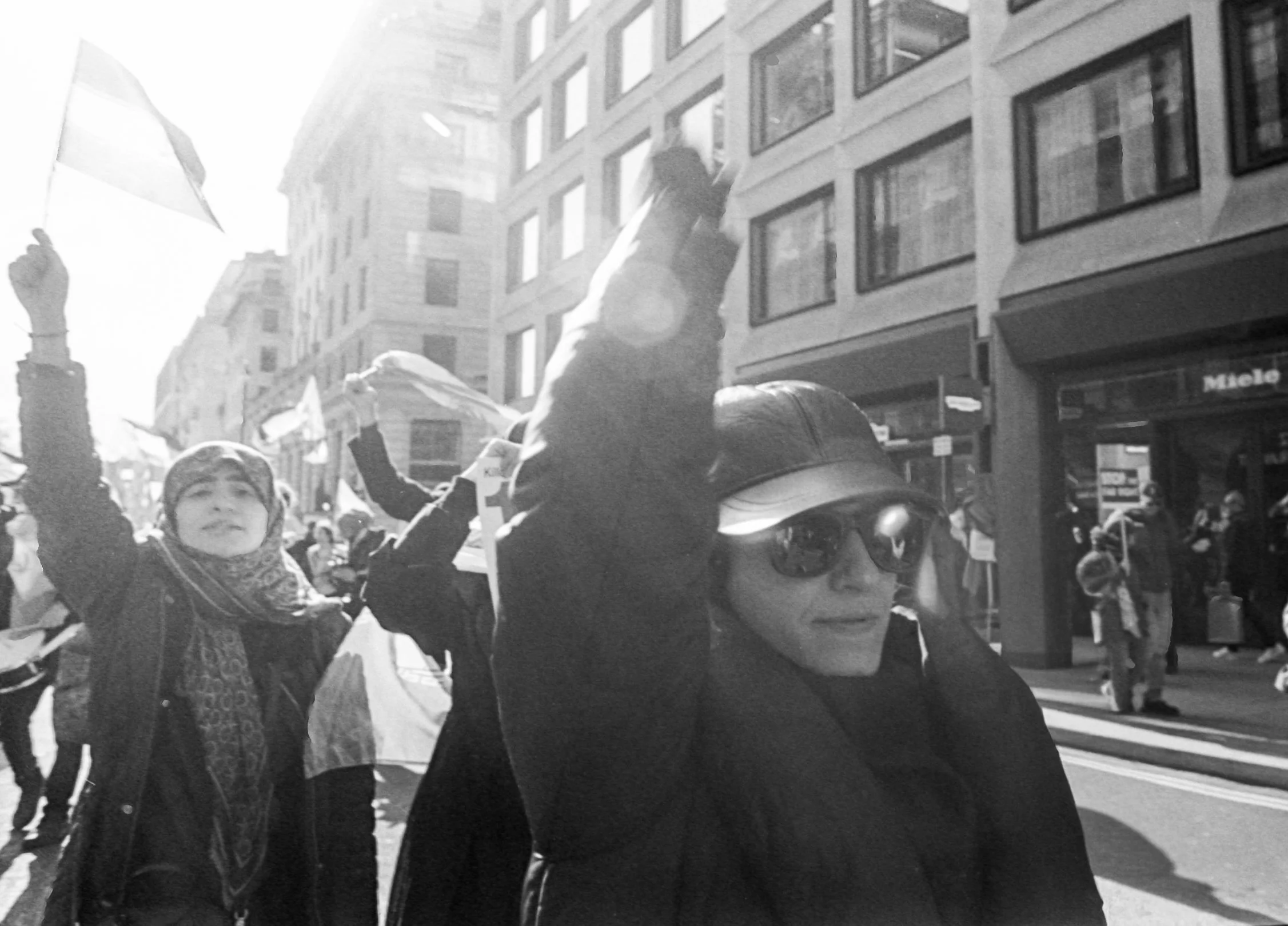 People participating in a street protest or demonstration, some holding flags, in an urban area with multi-story buildings.