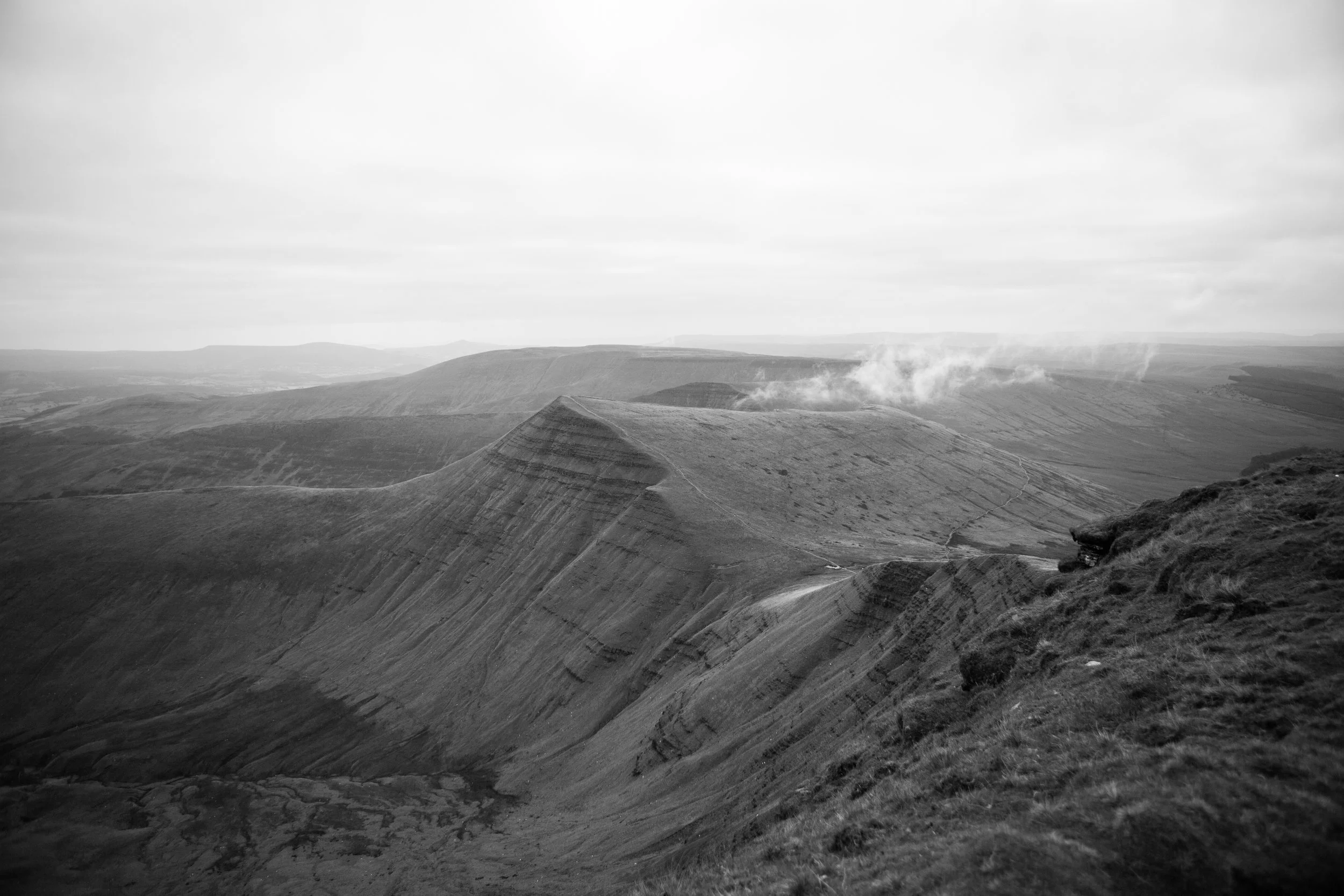 Black and white photo of rolling hills and ridges with patches of smoke or steam rising from the landscape, overcast sky in the background.