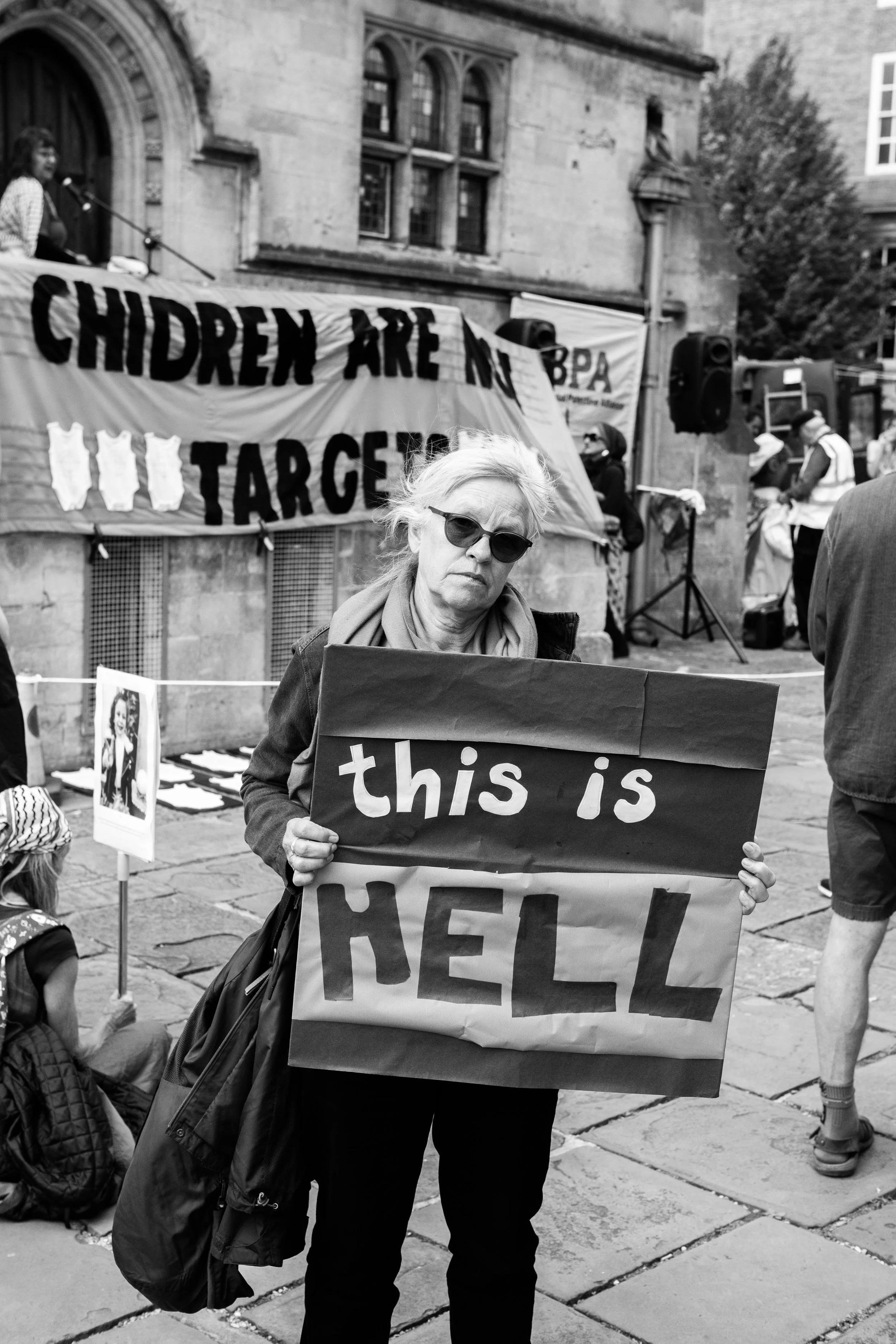 A woman with sunglasses holding a sign that says, 'this is HELL' at a protest or rally, with a banner in the background that reads 'CHILDREN ARE NOT TARGET'.