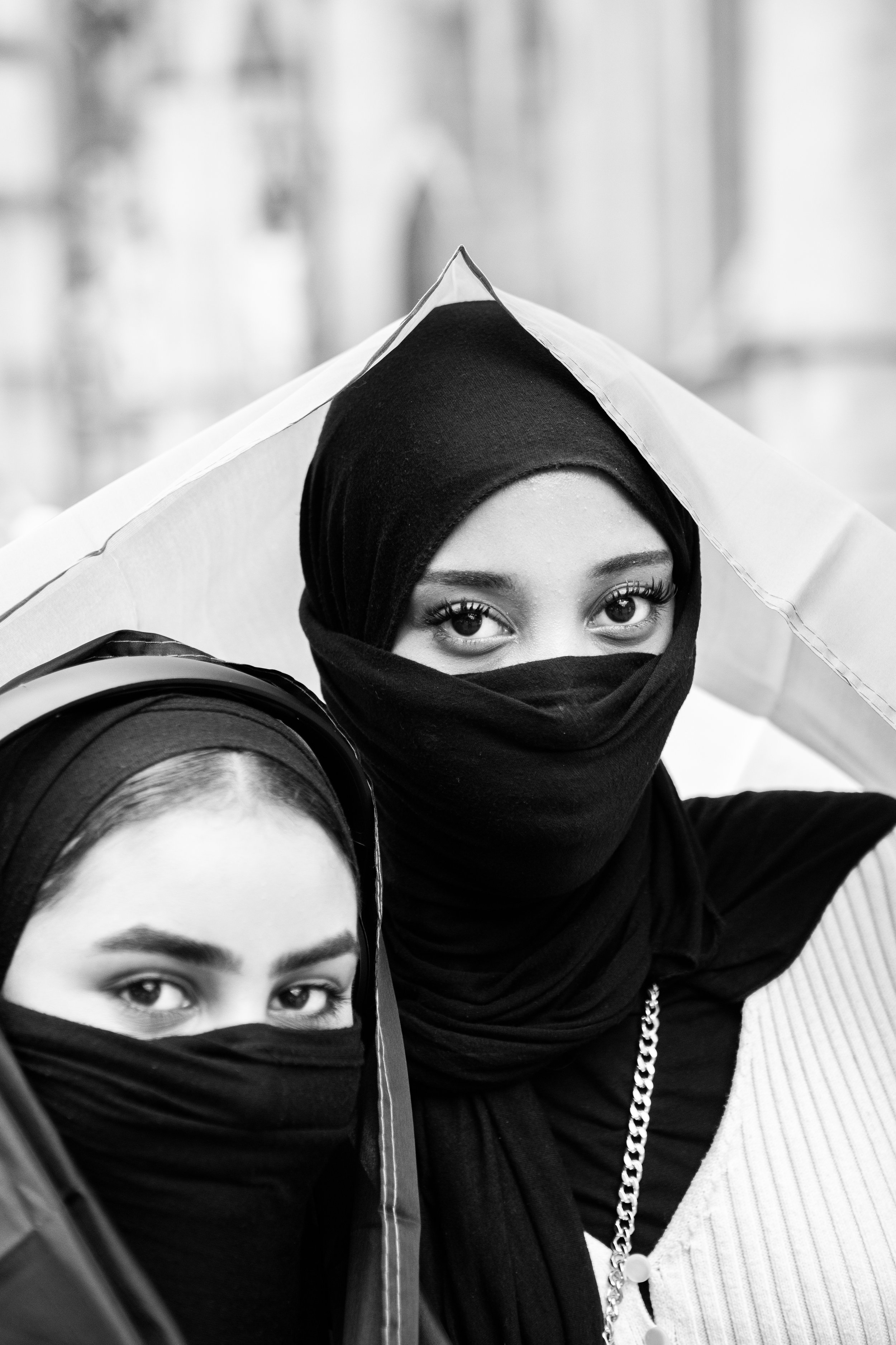 Two women wearing black hijabs and face coverings, standing closely together outdoors, with an umbrella overhead.