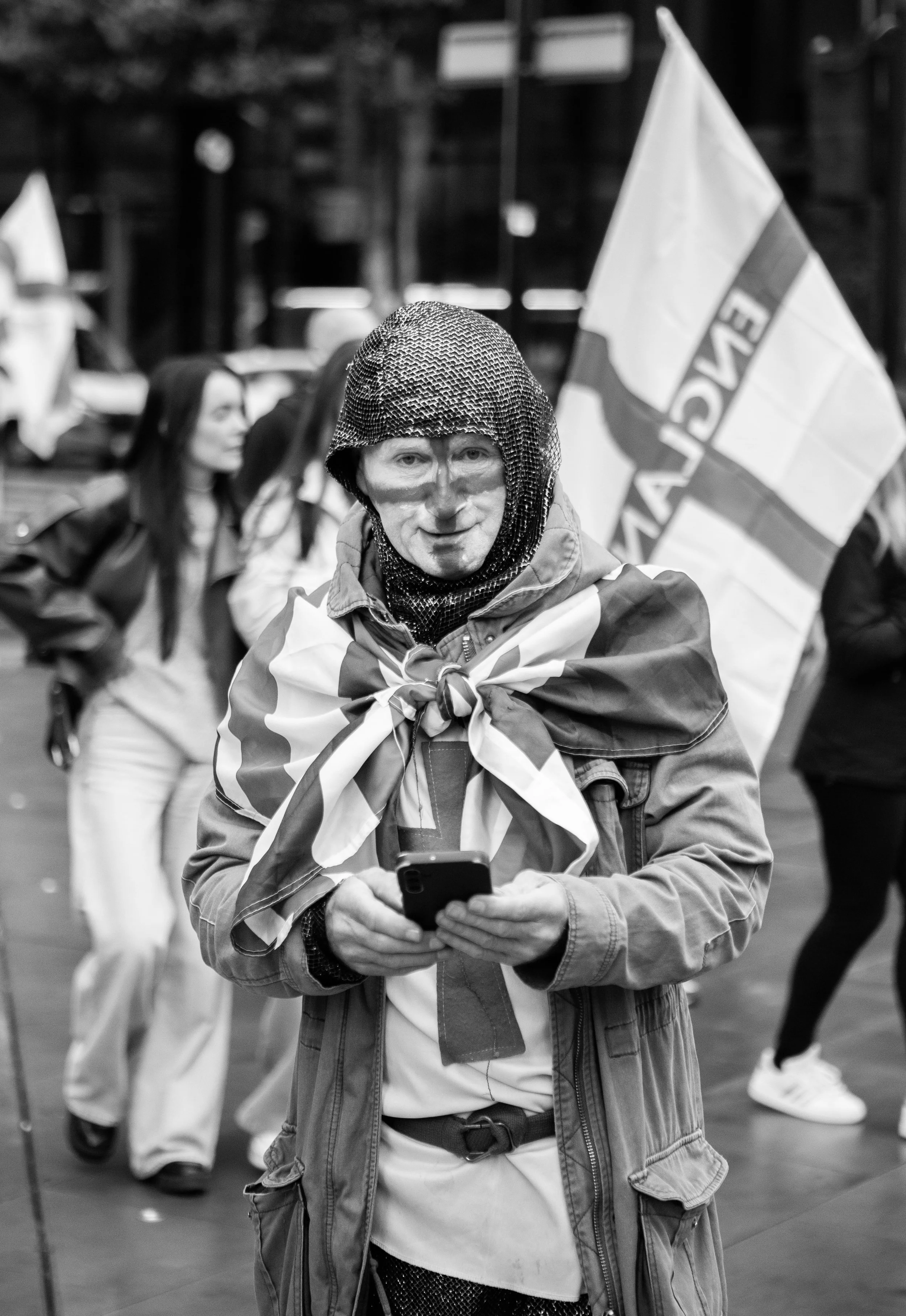 A person wearing a chainmail helmet and face paint, draped with a flag, holding a phone, at a protest or rally with other people and flags in the background.