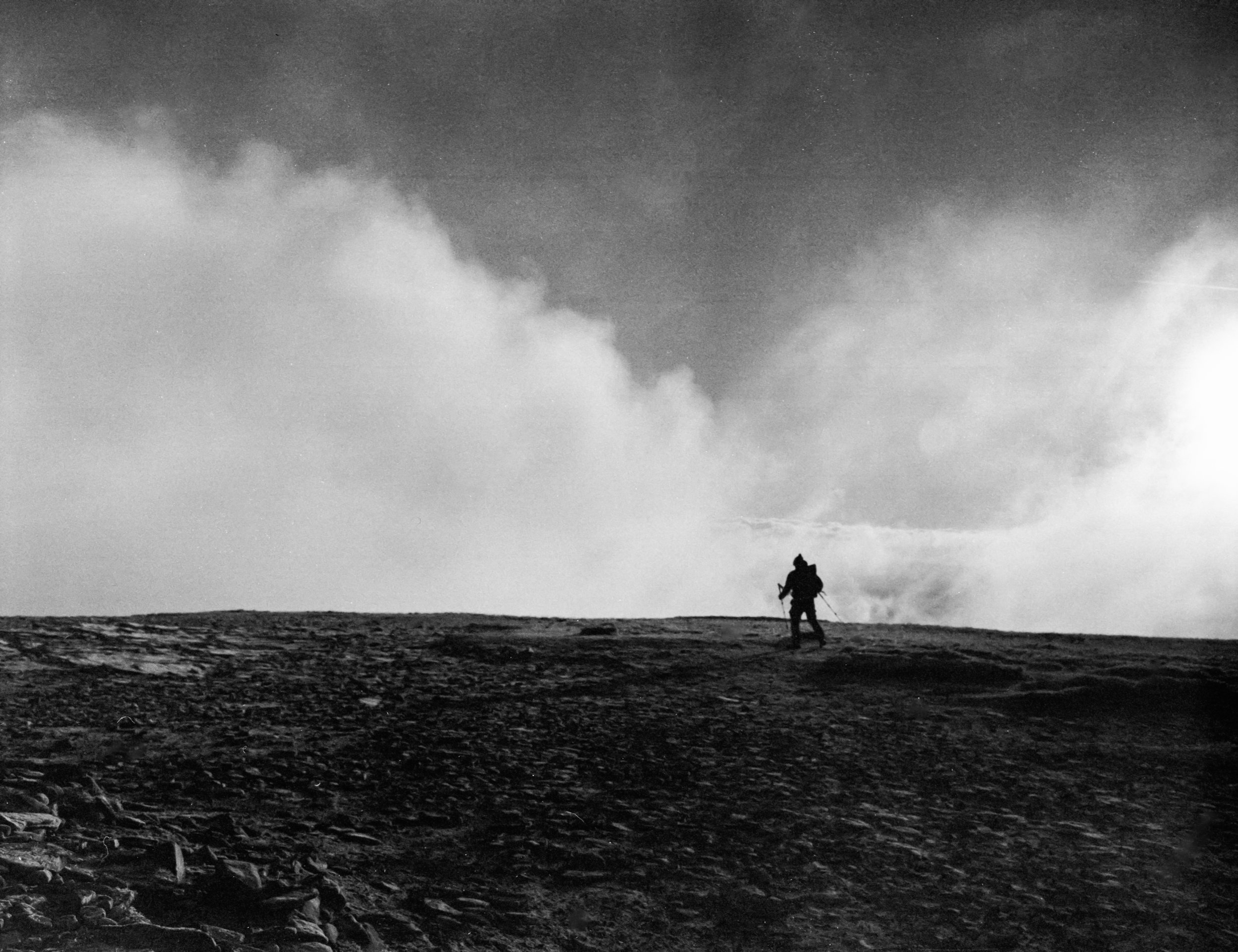 A lone hiker walking on a barren, rocky landscape with a cloudy sky overhead.