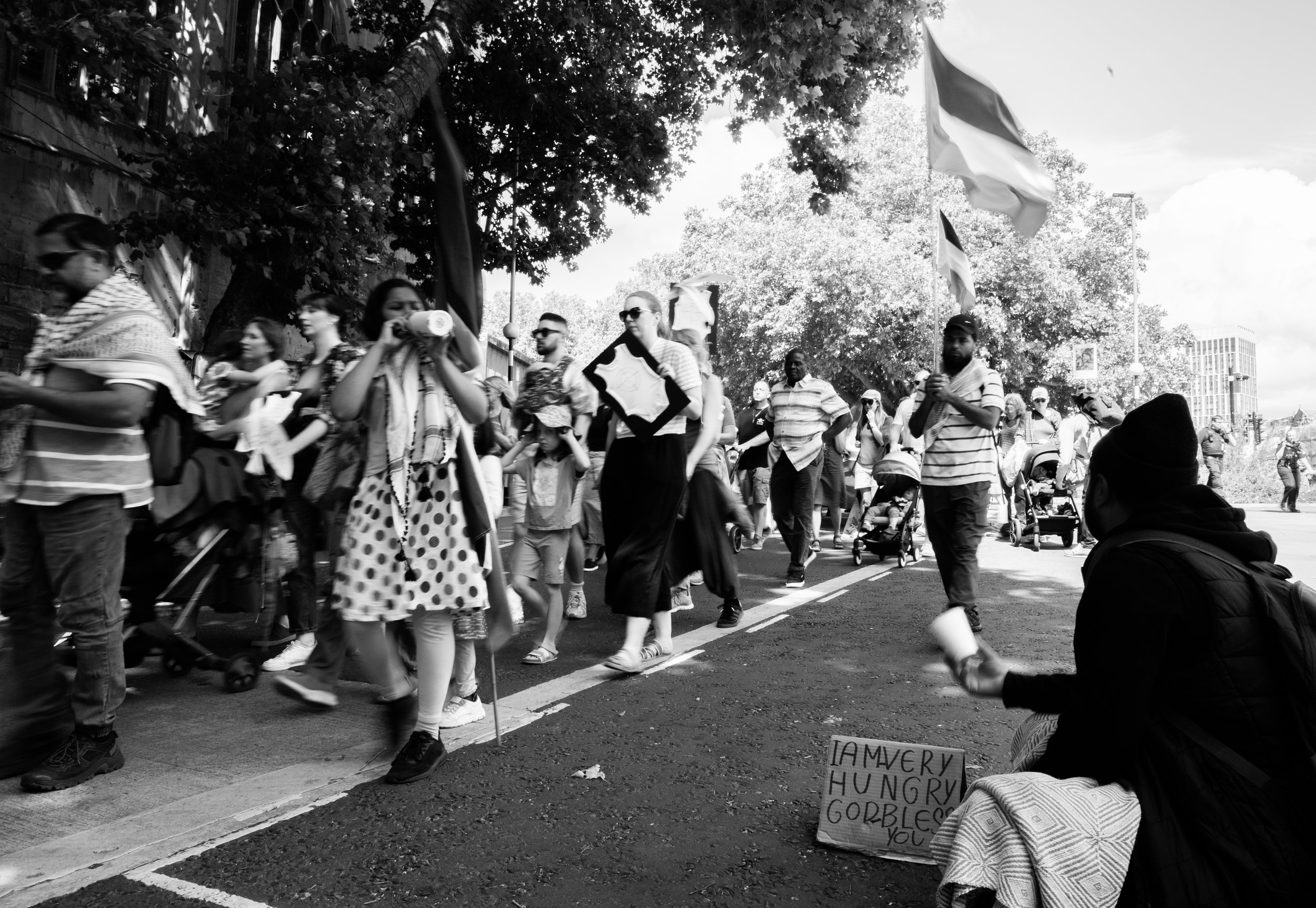 A black and white photo of a diverse group of people marching in a street parade with flags and signs, some pushing strollers and carrying framed items. A woman sitting on the sidewalk holds a handwritten sign that reads "I am very hungry goblet you.