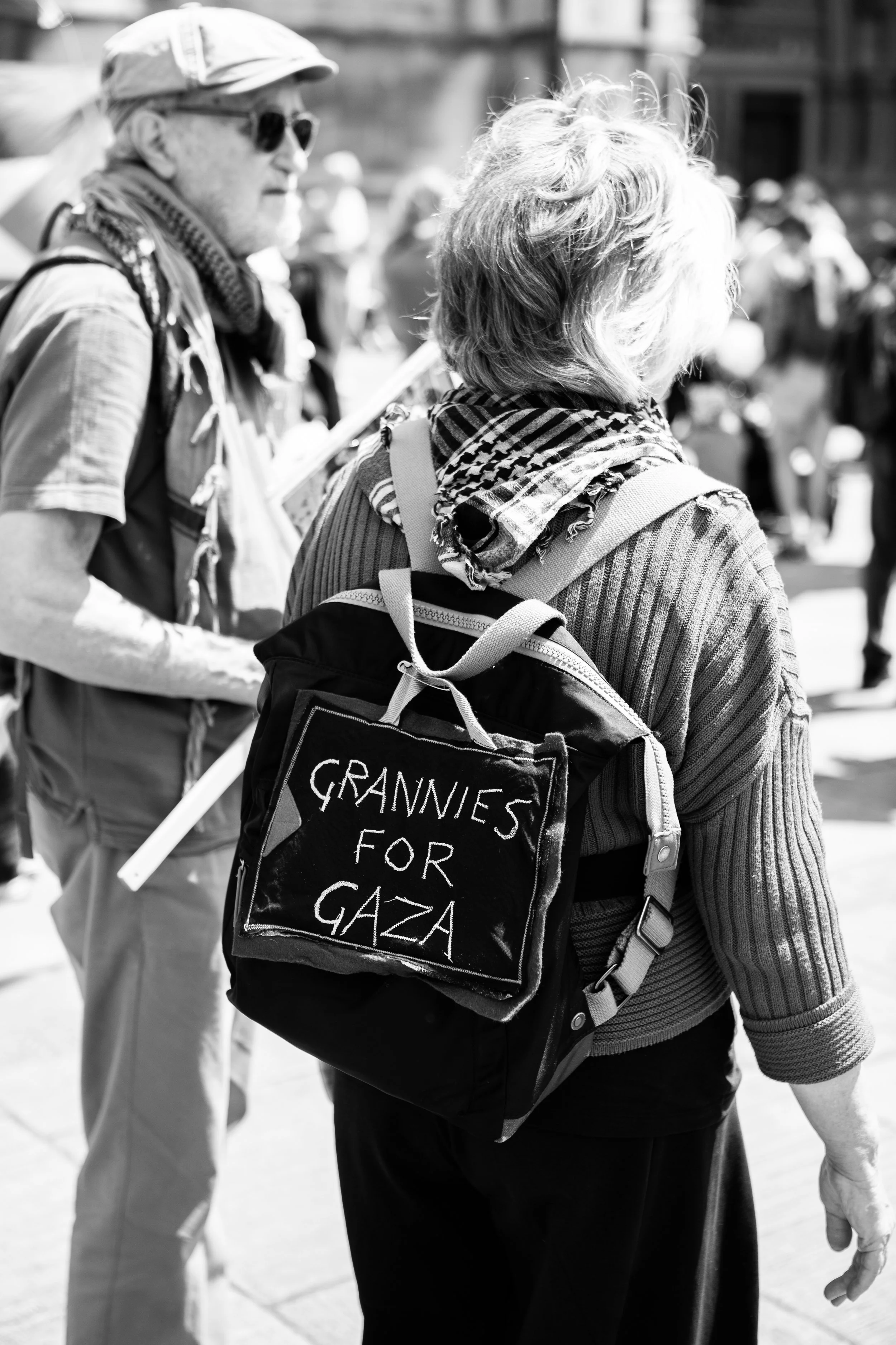 A woman with a backpack that has a sign saying 'Grannies for Gaza' visible on it, and a man holding a sign in the background, participating in a protest on a city street.