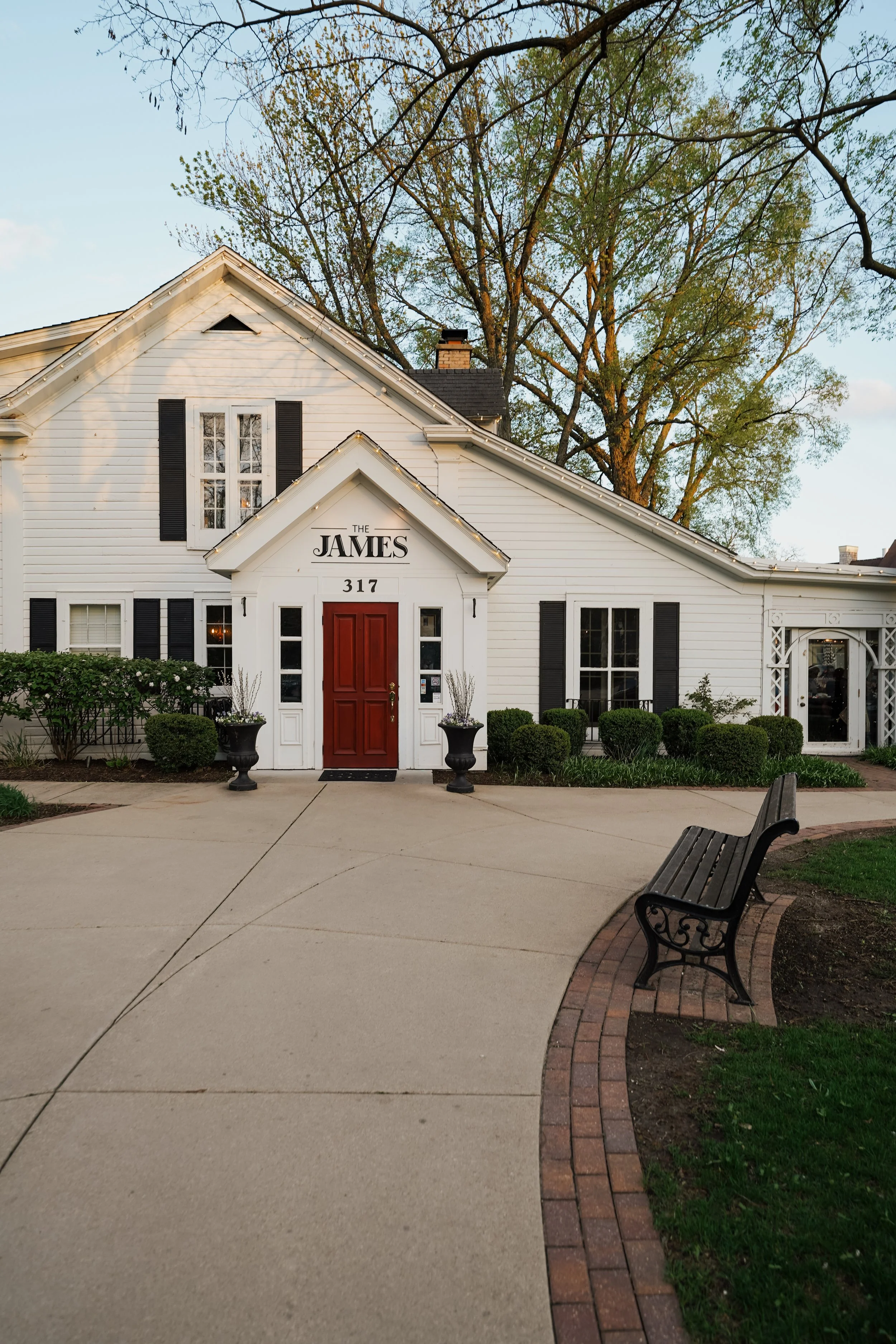 A white house with black shutters and a red door, labeled 'The James', 317, and surrounded by lawn, shrubs, and trees, is seen in the image. There is a curved sidewalk in front and benches on the grass.