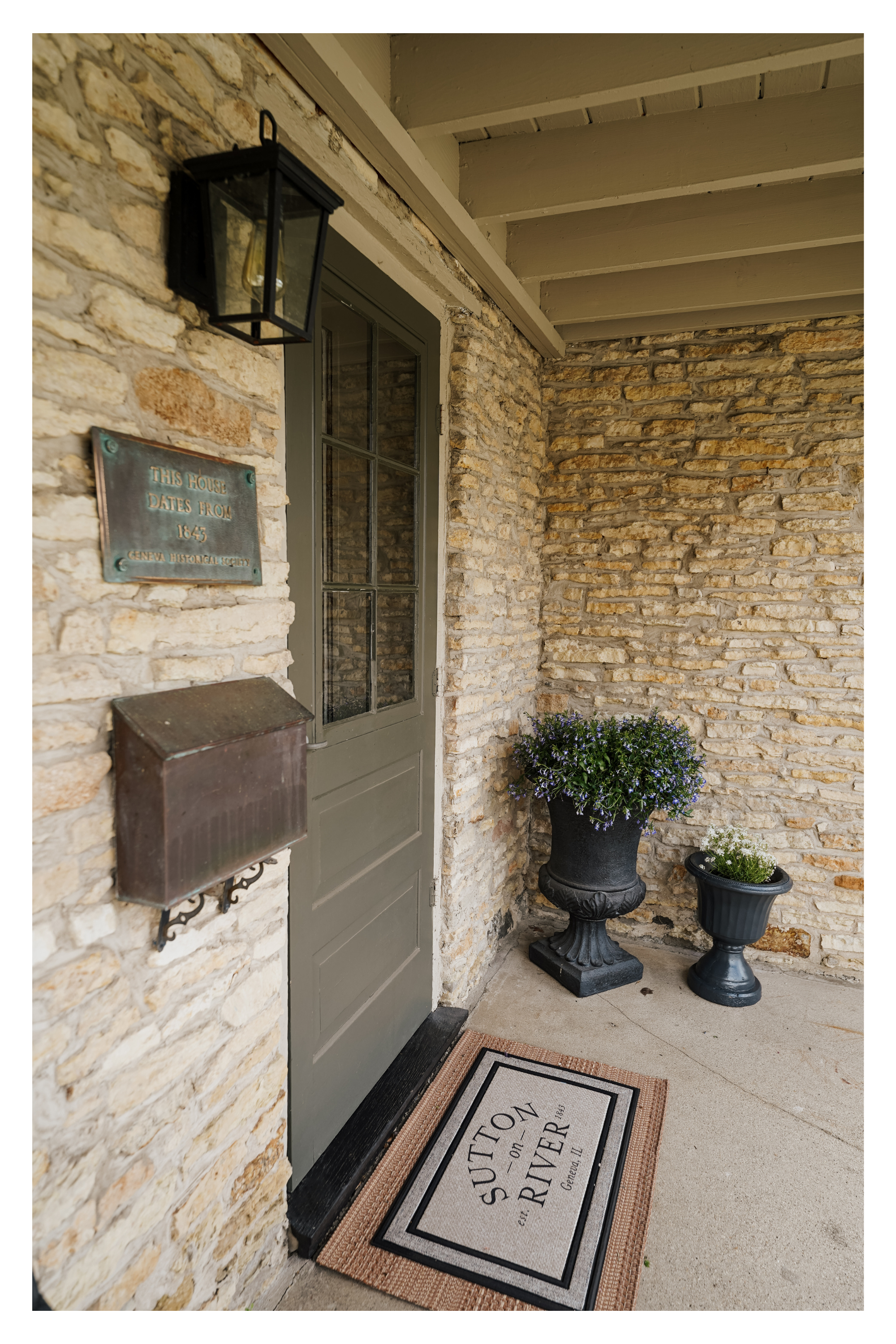 Stone house porch with a green door, black lantern light fixture, metal mailbox, two decorative plant pots with flowers, and a doormat reading "SUTTON ON THE RIVER," with a brick wall and an overhanging roof.