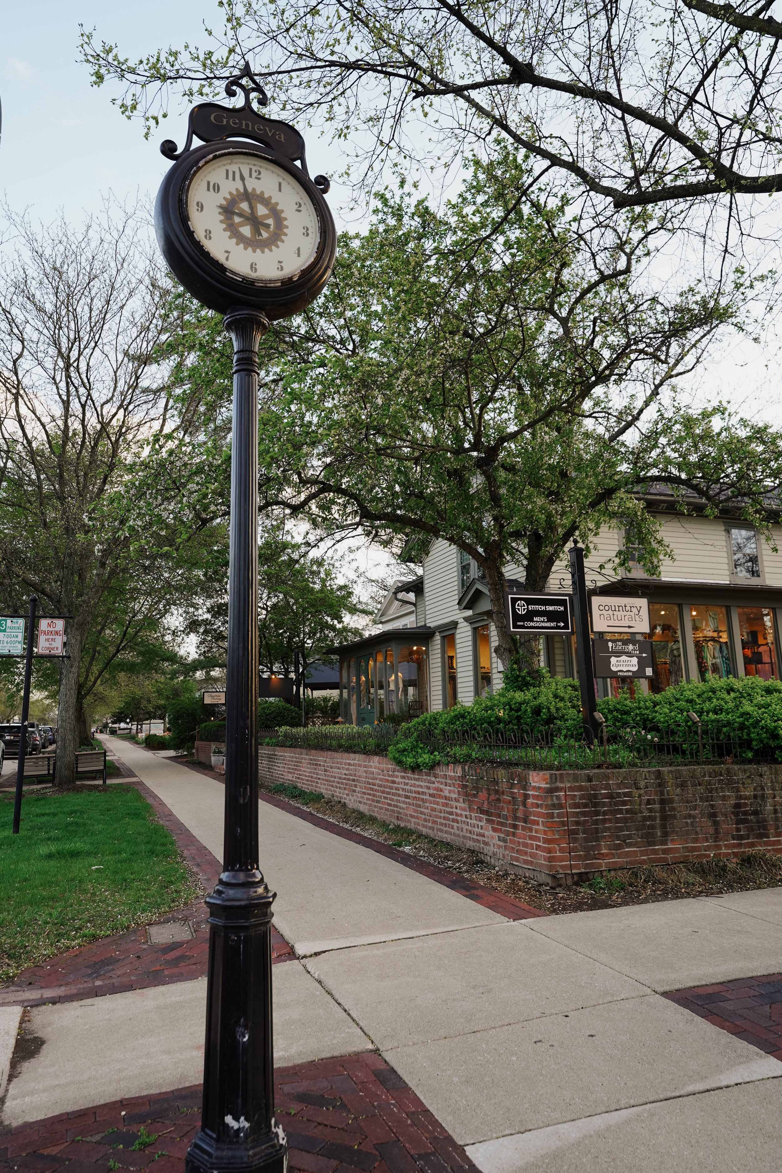 A black street clock on a tall pole labeled 'Geneva', with trees and a building with shop signs in the background on a sidewalk in Geneva, Illinois.