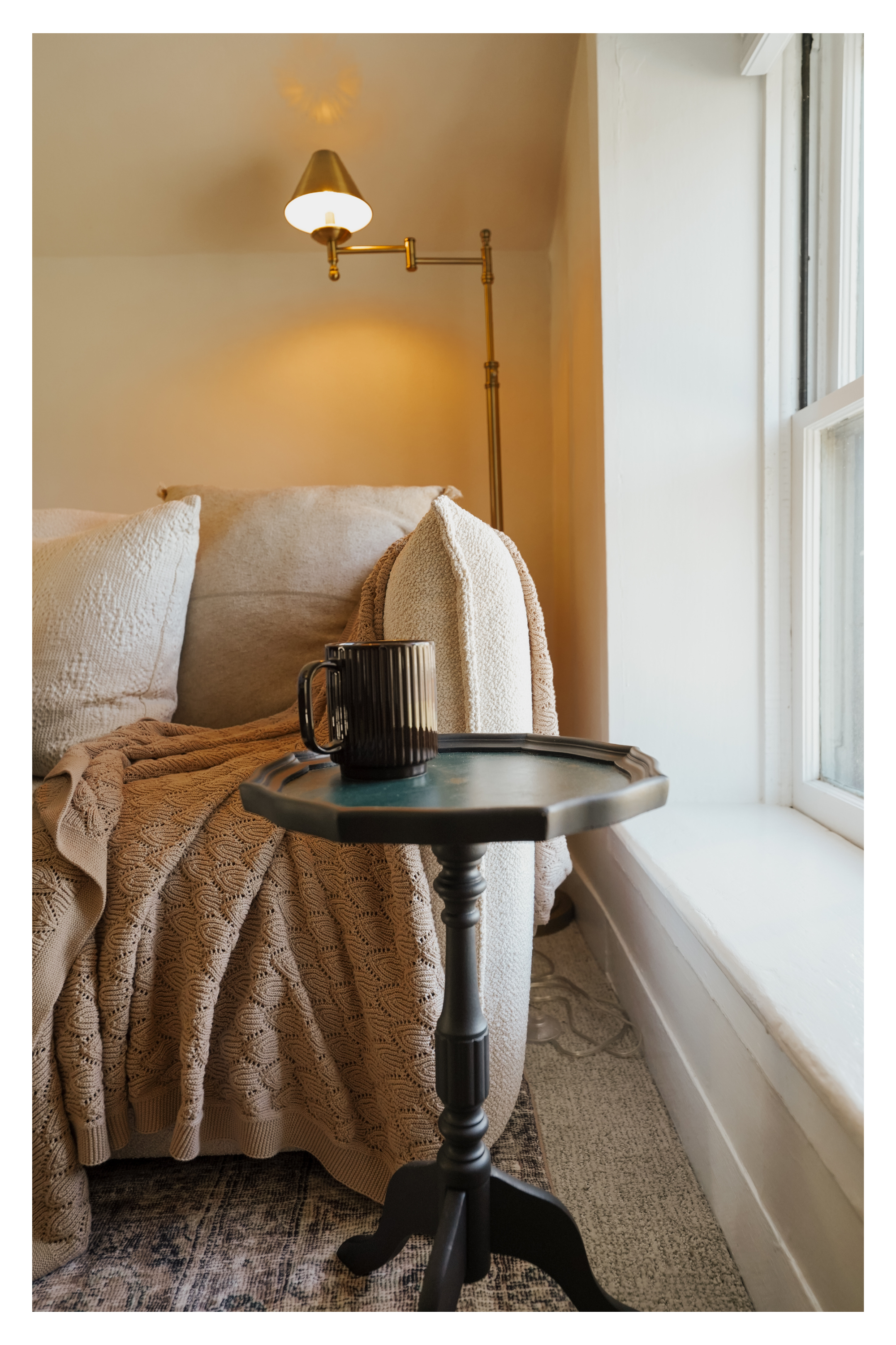 A cozy reading nook with a beige sofa, textured throw pillows, a knitted blanket, a small round side table with a black mug, a vintage-style adjustable reading lamp, a window with white trim, and a patterned area rug.