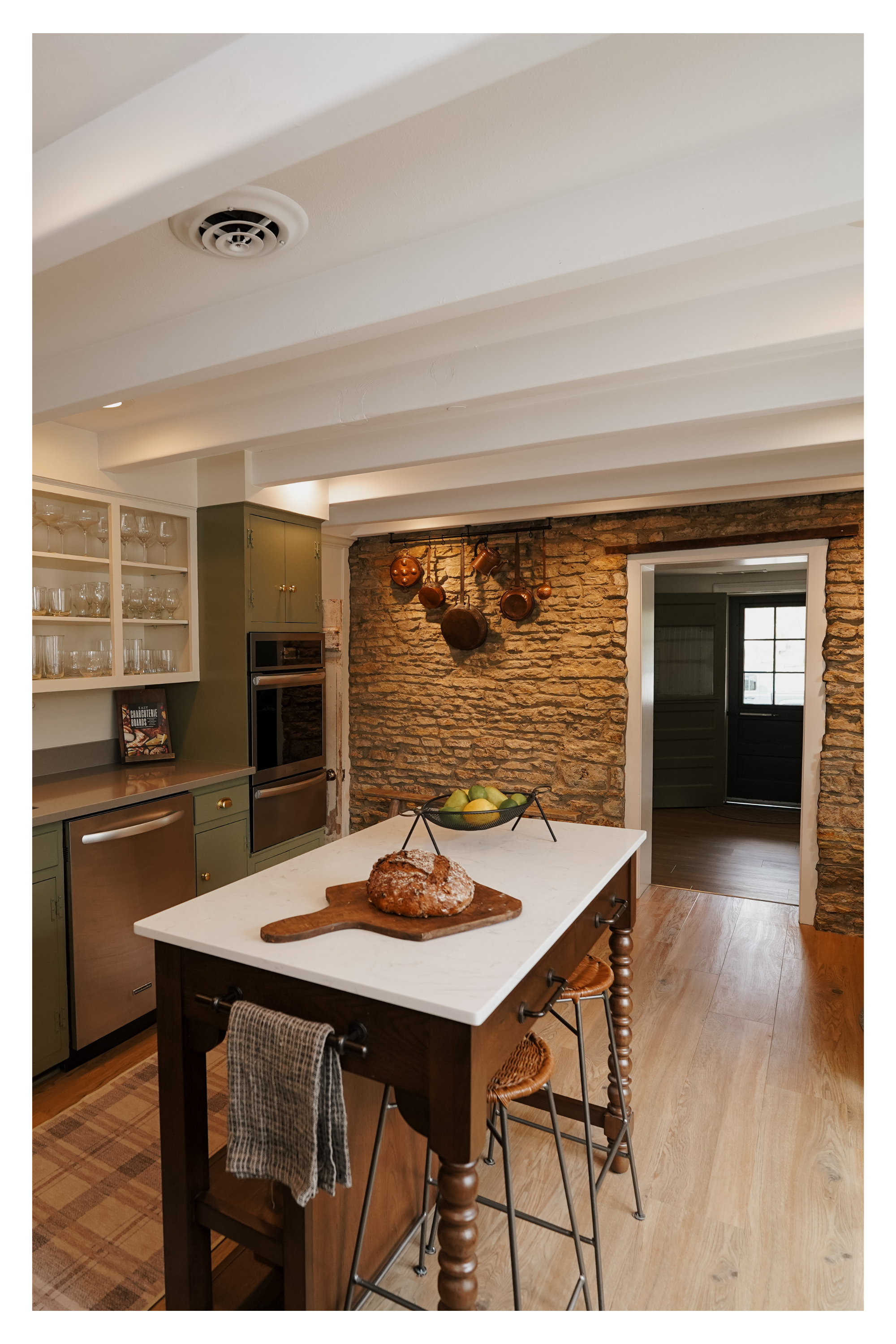 A rustic kitchen with exposed brick wall, white ceiling beams, and a kitchen island with a loaf of bread on a cutting board, surrounded by metal stools, green cabinets, and a hanging pot rack.