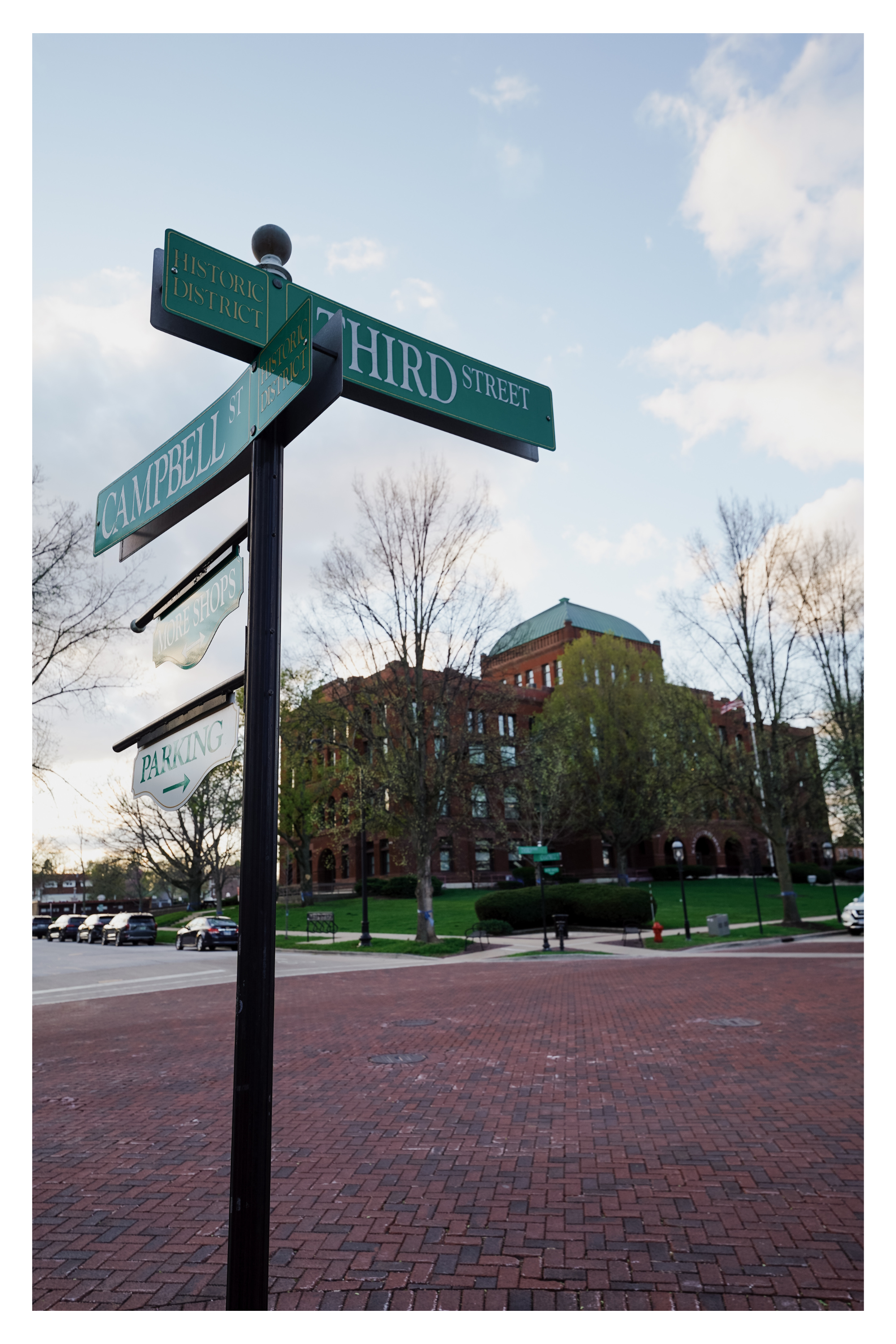 Street corner with street signs indicating Third Street and Campbell Street in a Geneva, Illinoihistoric district, with a brick-paved sidewalk, parked cars, a park area with trees, benches, and a historic building with a green dome in the background.