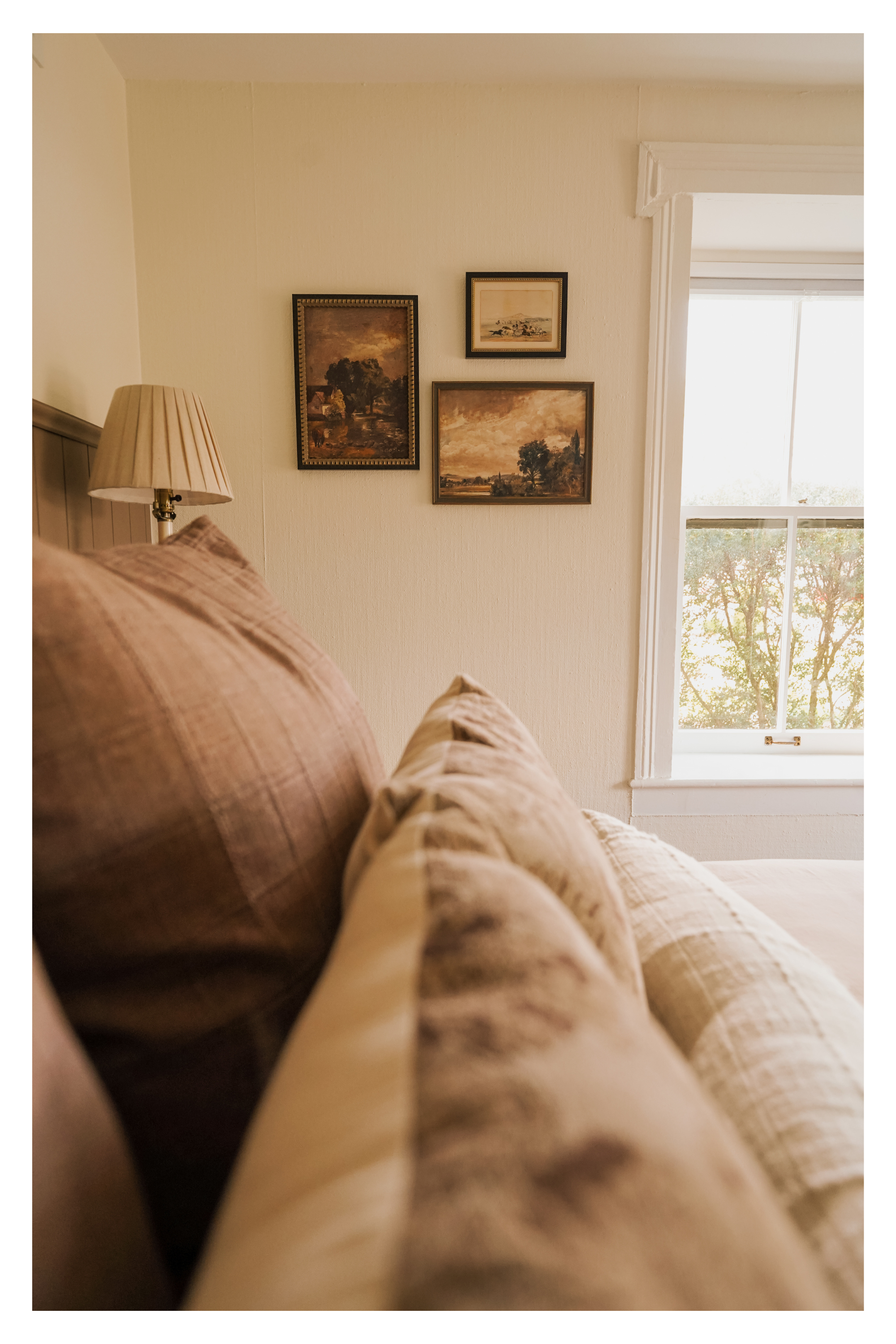 A cozy bedroom corner with pillows on a bed, a lamp, and framed landscape paintings on the wall next to a window with white trim showing trees outside.