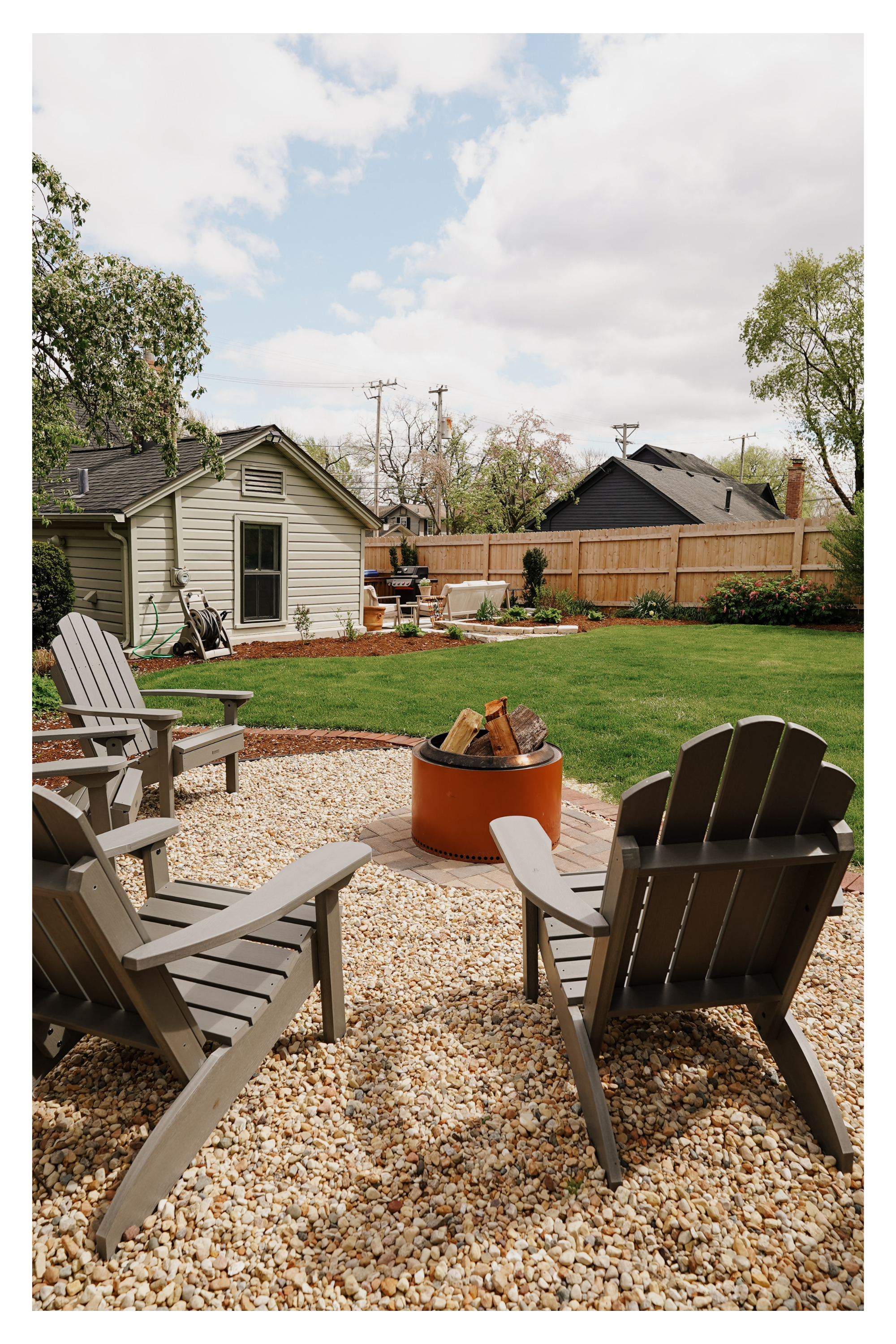 Backyard patio with Adirondack chairs around a fire pit, a shed, and a lawn, with a wooden fence and trees in the background.