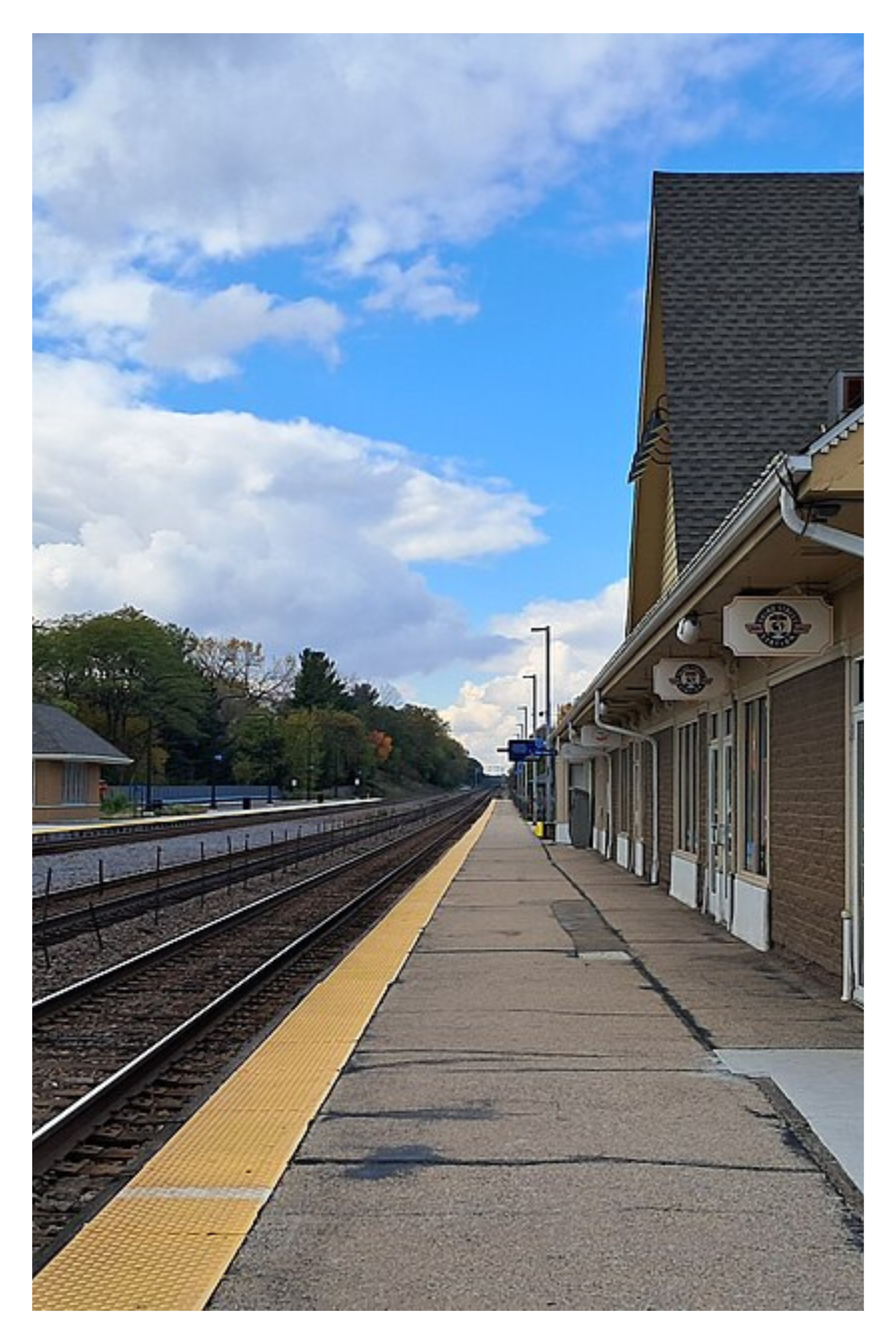 Empty train station platform with train tracks, yellow tactile paving edge, and a building with signage. Trees and a partly cloudy sky are visible in the background.