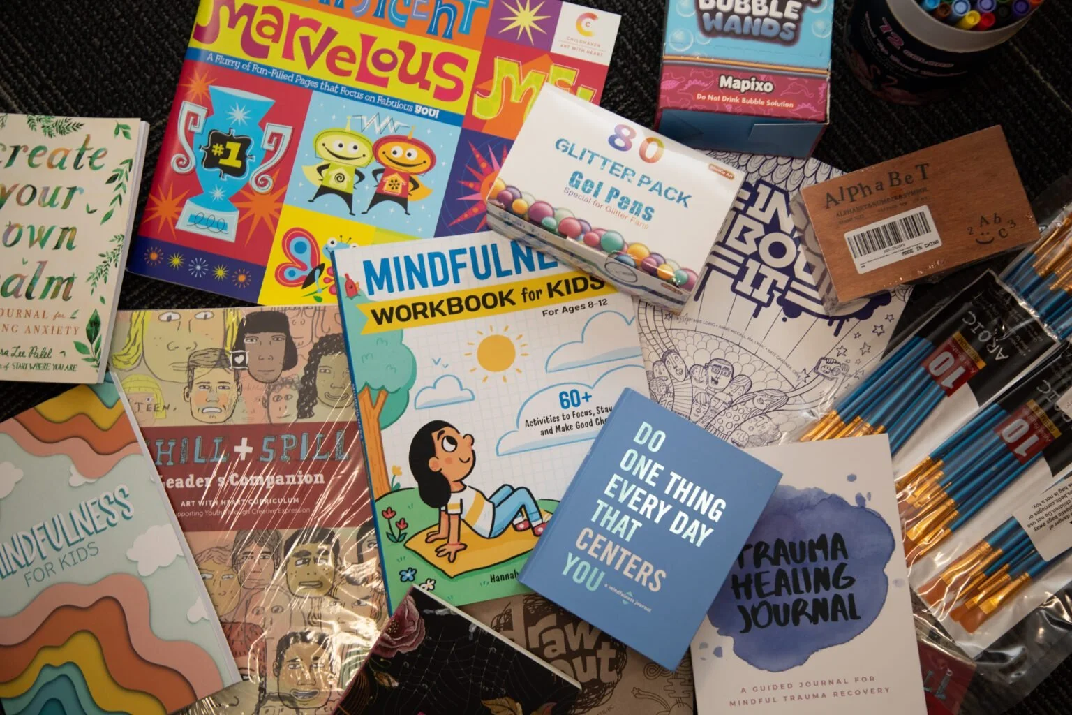 A variety of colorful books, workbooks, journals, and stationery items spread out on a dark surface, including books on mindfulness for kids, a glitter pack, a comic book, and pens.