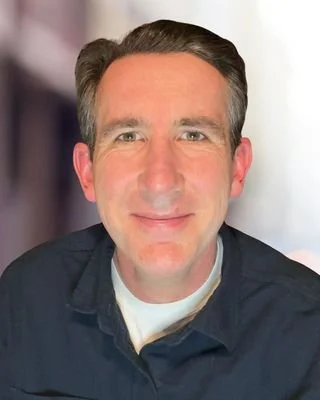 A man with short dark hair, wearing a dark shirt and smiling at the camera.