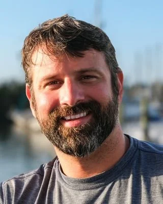A beige ceramic vase with dried branches on a wooden surface, casting a shadow on a plain wall.