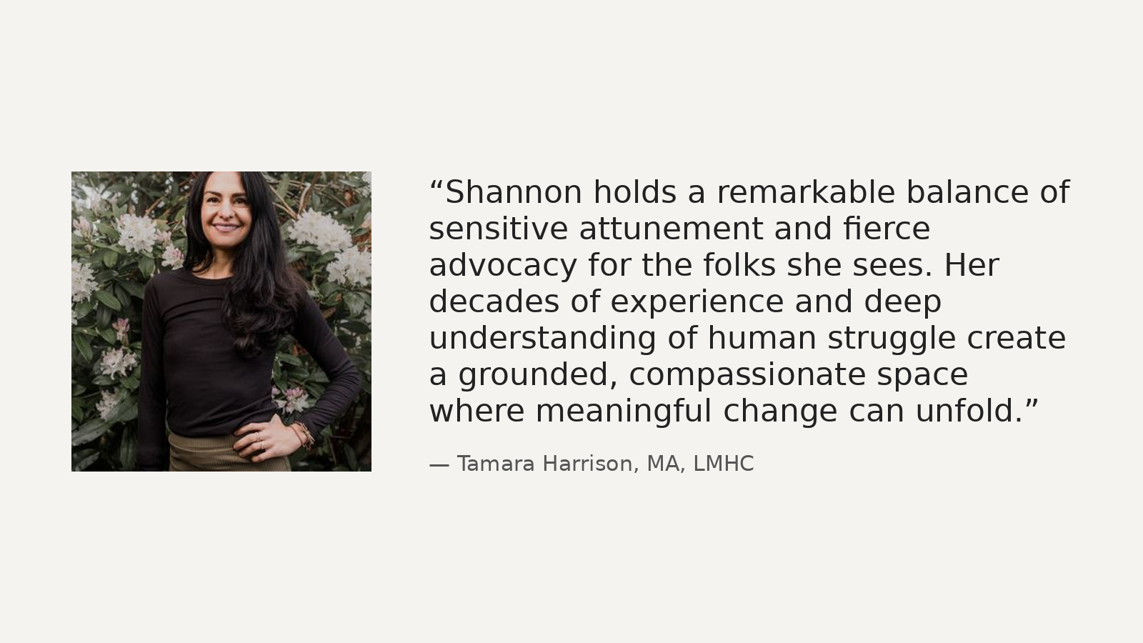 Photo of a woman with long dark hair wearing a black long-sleeve shirt, standing outdoors in front of flowering shrubbery, smiling with her hand on her hip.