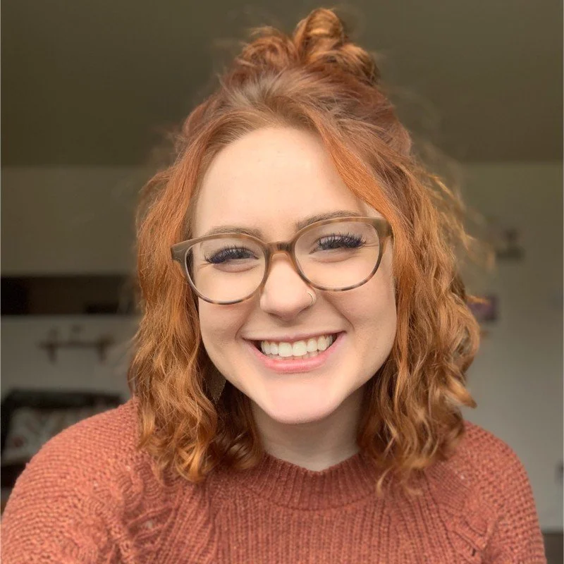 A young woman with red curly hair and glasses smiling at the camera.