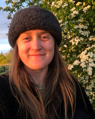 A beige ceramic vase with a simple, curved shape holding a small dry branch, placed on a wooden surface with sunlight creating shadows in the background.