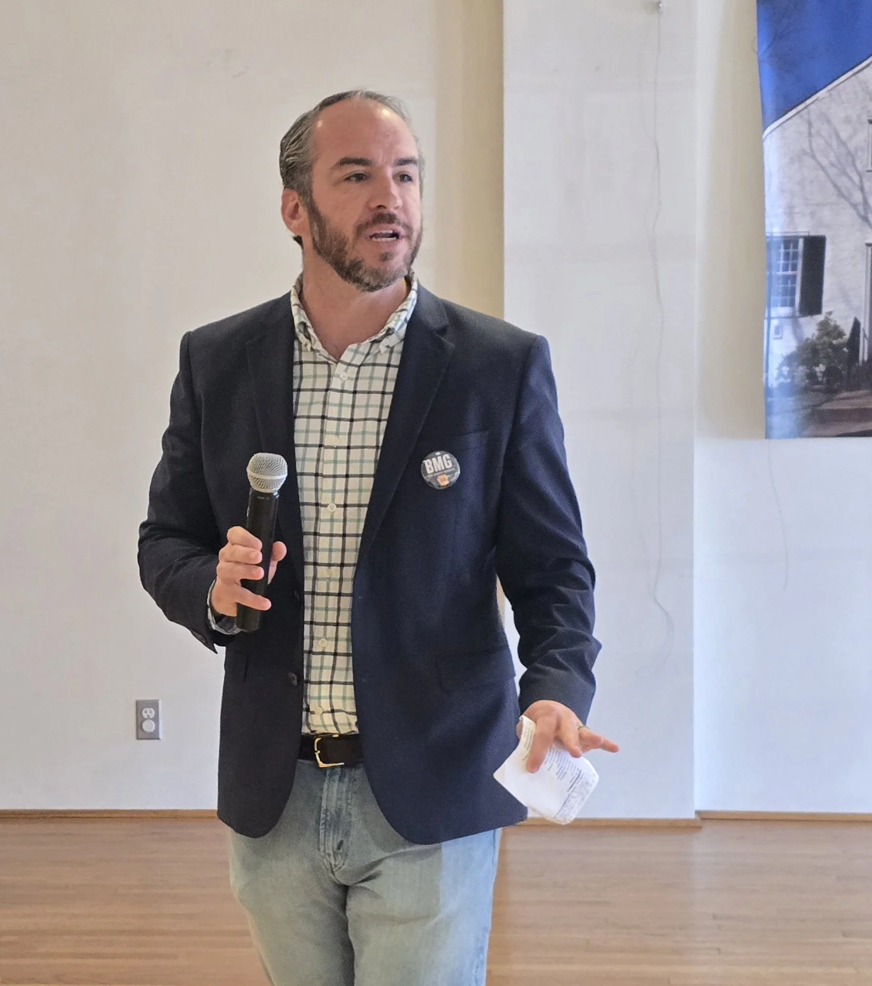 Bradley Merkl-Gump wearing a dark blazer, plaid shirt, and jeans holding a microphone and a piece of paper, speaking at an indoor event.