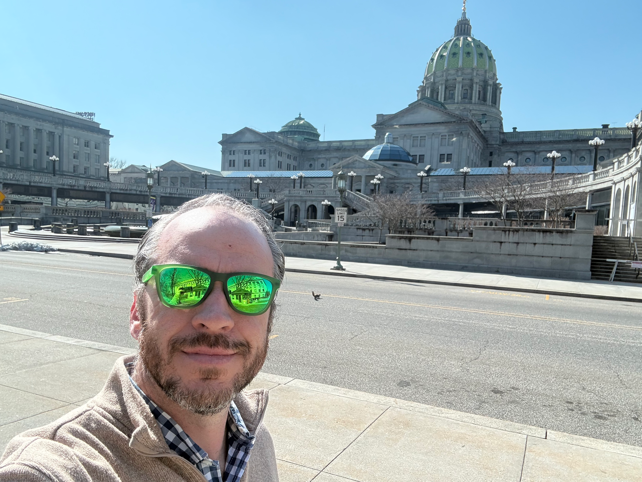 Bradley Merkl-Gump, candidate for Pennsylvania senate, wearing sunglasses in front of Harrisburg capitol building with a dome, stairs, and columns, under a clear blue sky.