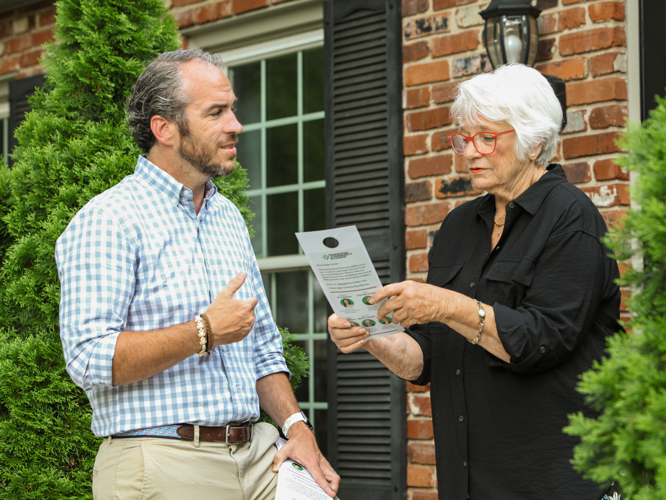A man and an older woman standing outside a brick house engaged in conversation. The woman is holding a pamphlet and explaining something to the man, who is pointing at her. Green bushes surround them.