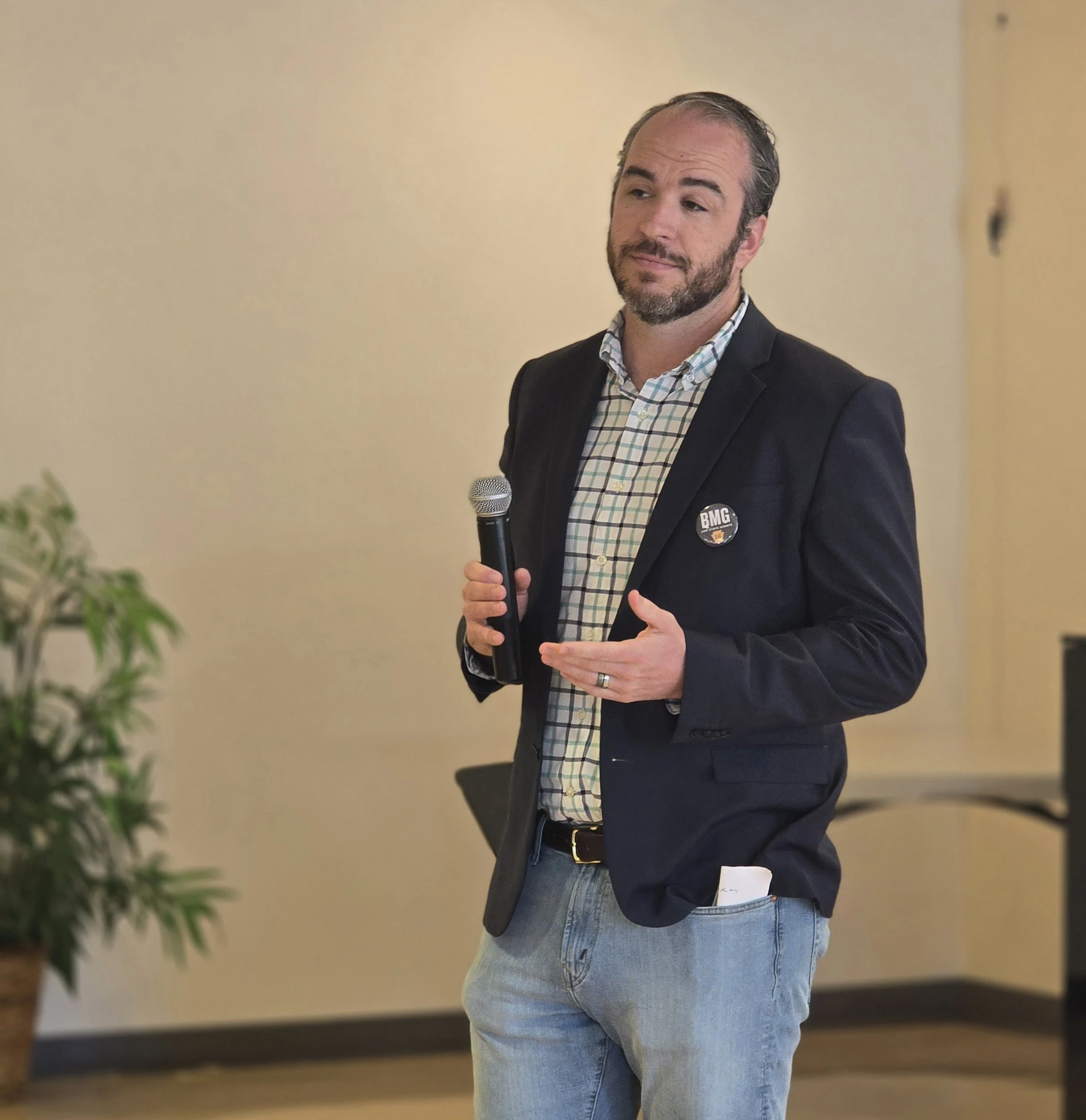 Bradley Merkl-Gump in a black blazer, plaid shirt, and jeans holding a microphone, standing in a room with a potted plant, giving a speech.