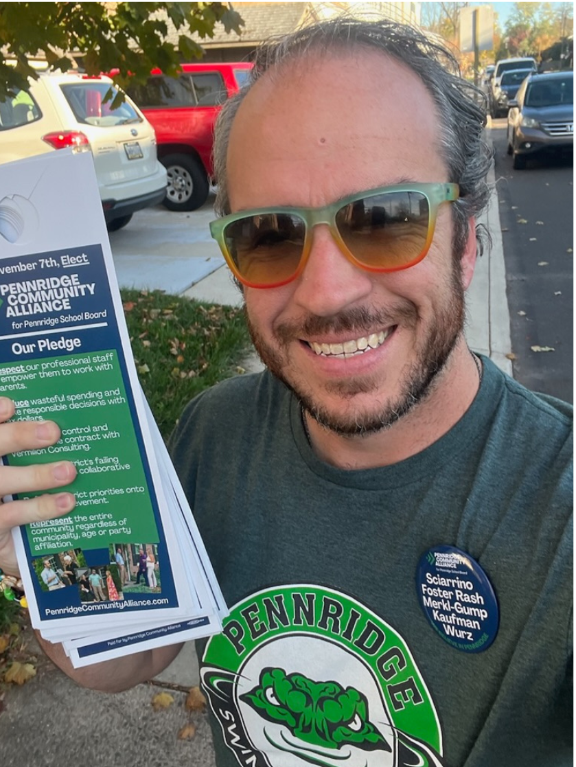 Bradley Merkl-Gump with sunglasses smiling and holding pamphlets at a community event. He is wearing a Pennridge shirt and a button with names of candidates.